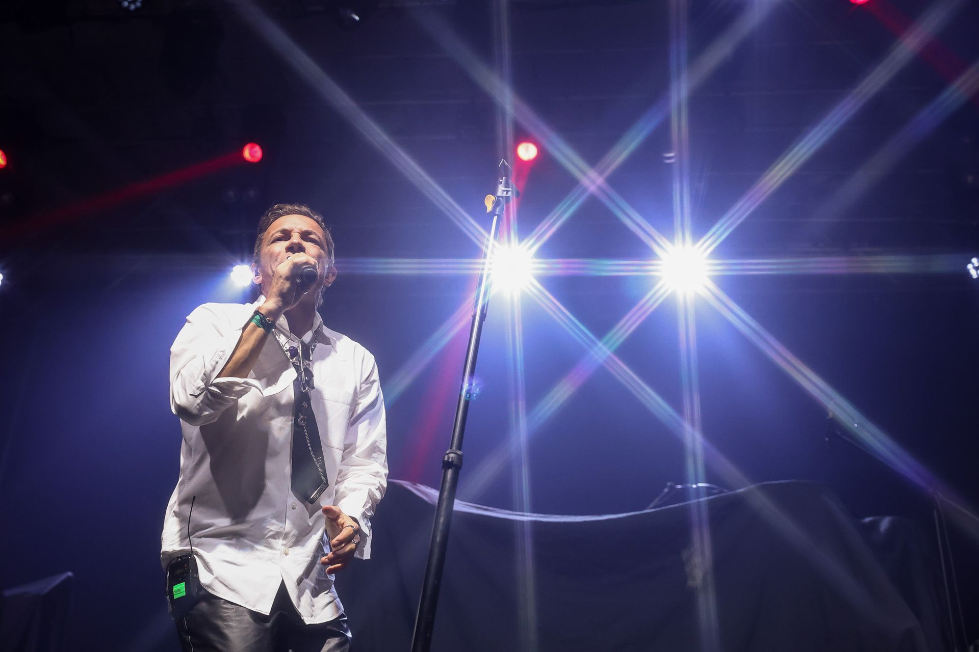 Um artista de camisa branca e gravata canta em um microfone em um palco iluminado por holofotes brilhantes