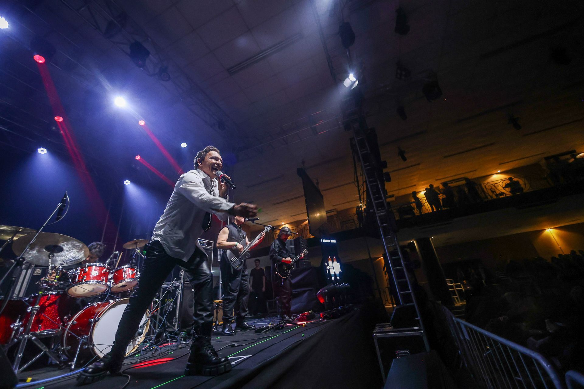 Uma cantor de camisa branca se apresenta no palco com uma banda sob luzes de palco azuis e brancas em um show.
