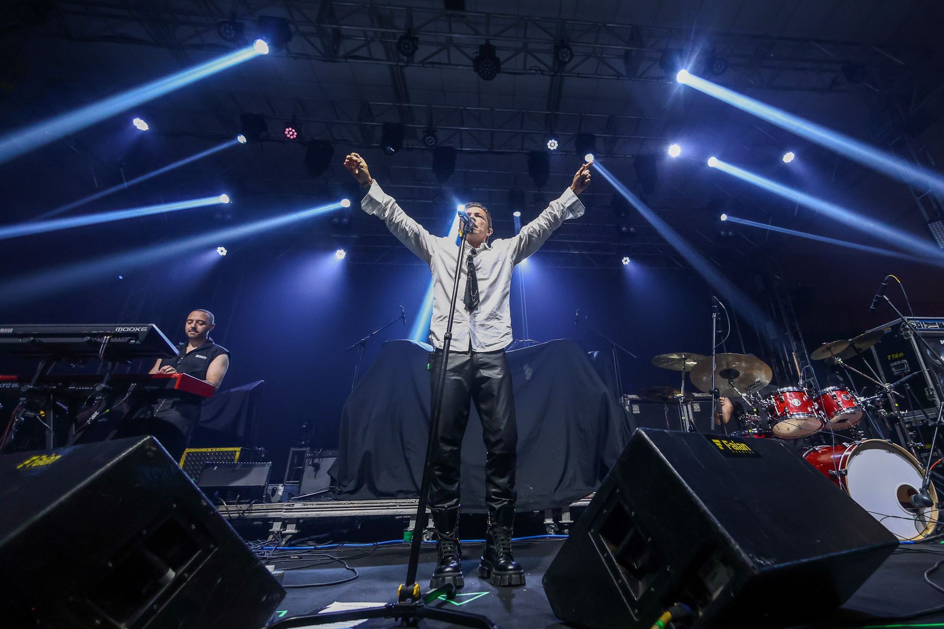Um artista está no centro do palco com os braços erguidos, cercado por luzes de concerto e equipamentos de banda.