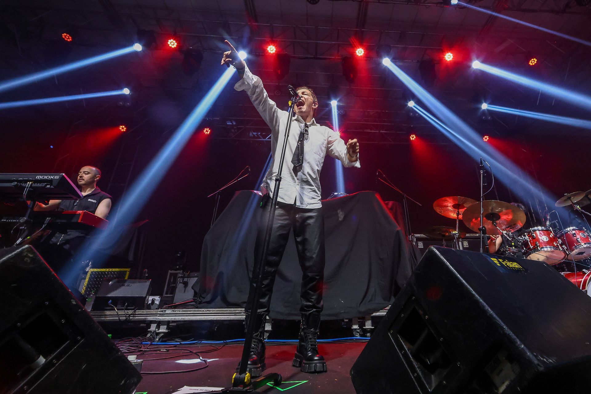 Um artista de camisa branca e gravata canta em um microfone no palco, com uma banda e luzes brilhantes de estádio atrás dele.