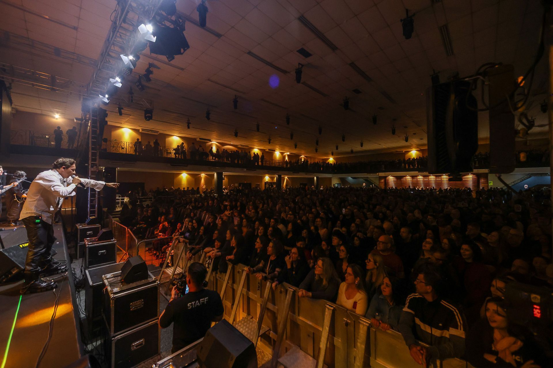 Um músico se apresenta no palco com um microfone diante de uma grande plateia em um local fechado com pouca luz.