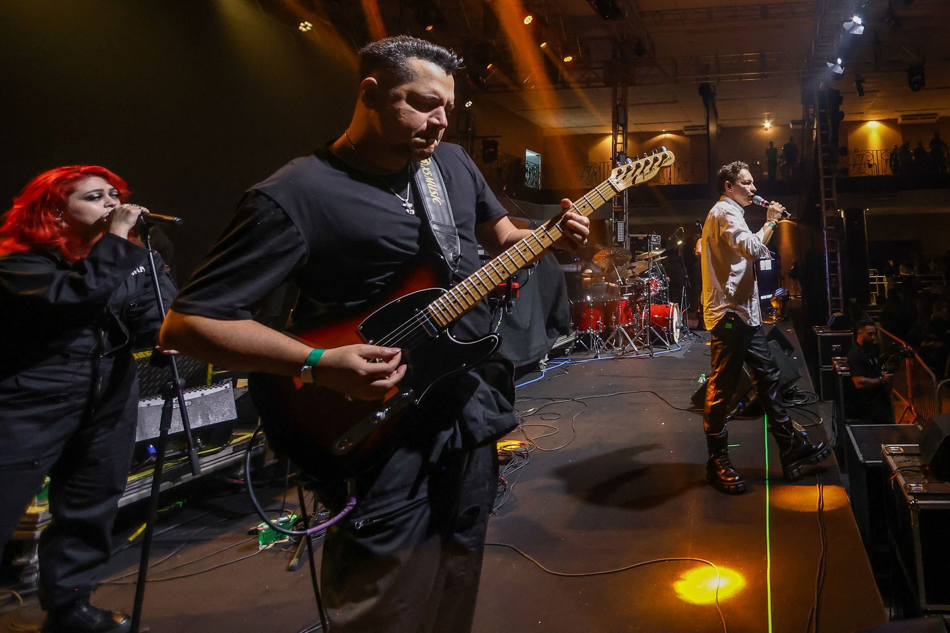 Uma banda se apresenta no palco: um guitarrista toca, uma pessoa ruiva canta ao microfone e um vocalista está à direita.