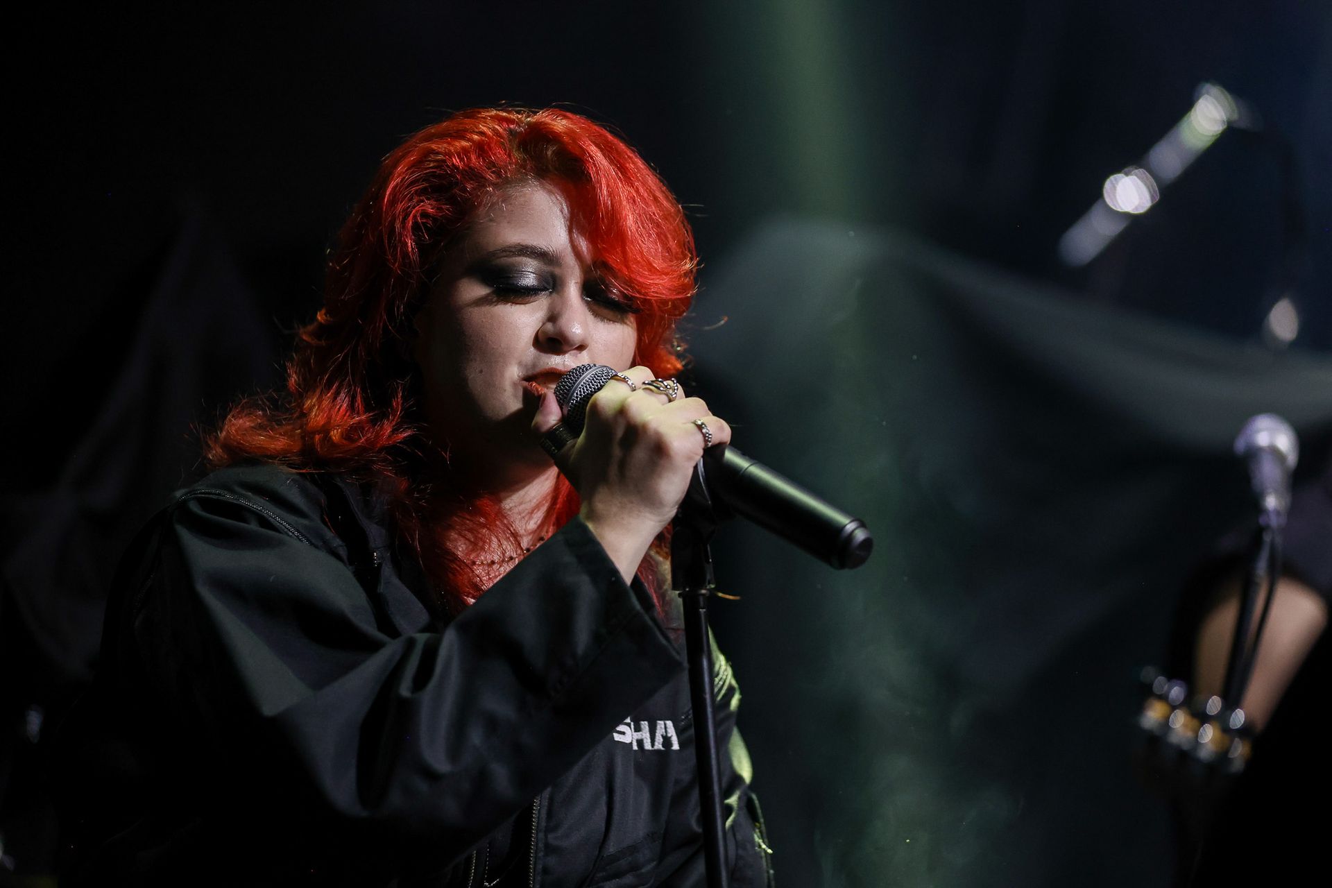 Uma pessoa com cabelos ruivos vibrantes cantando em um microfone em um palco pouco iluminado.
