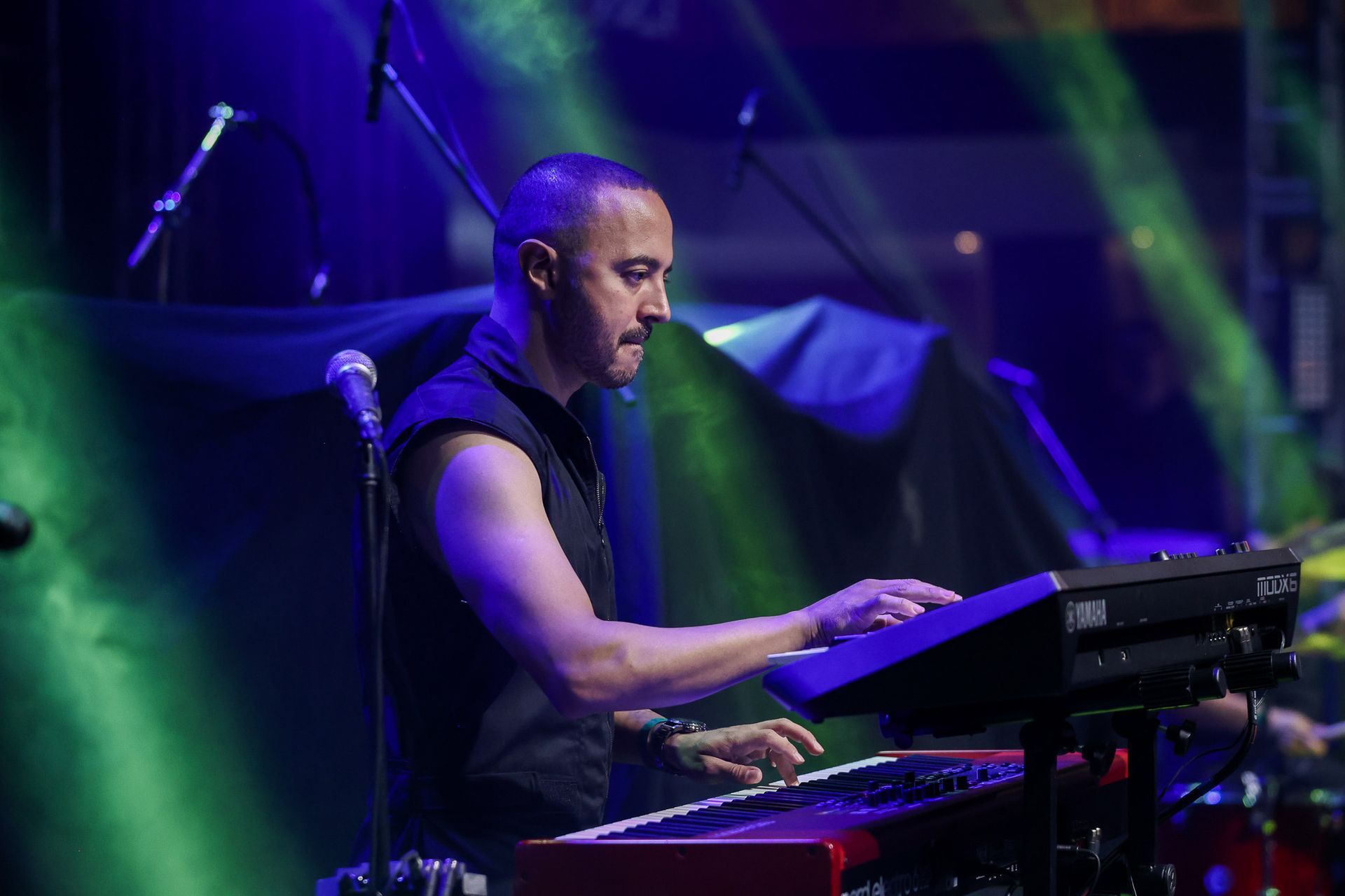 Um músico toca teclado em um palco pouco iluminado por luzes de concerto azuis e verdes.