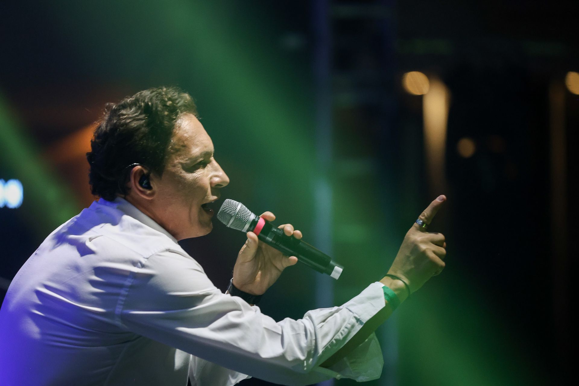 Uma pessoa vestindo uma camisa branca cantando em um microfone no palco, apontando para cima com uma das mãos.