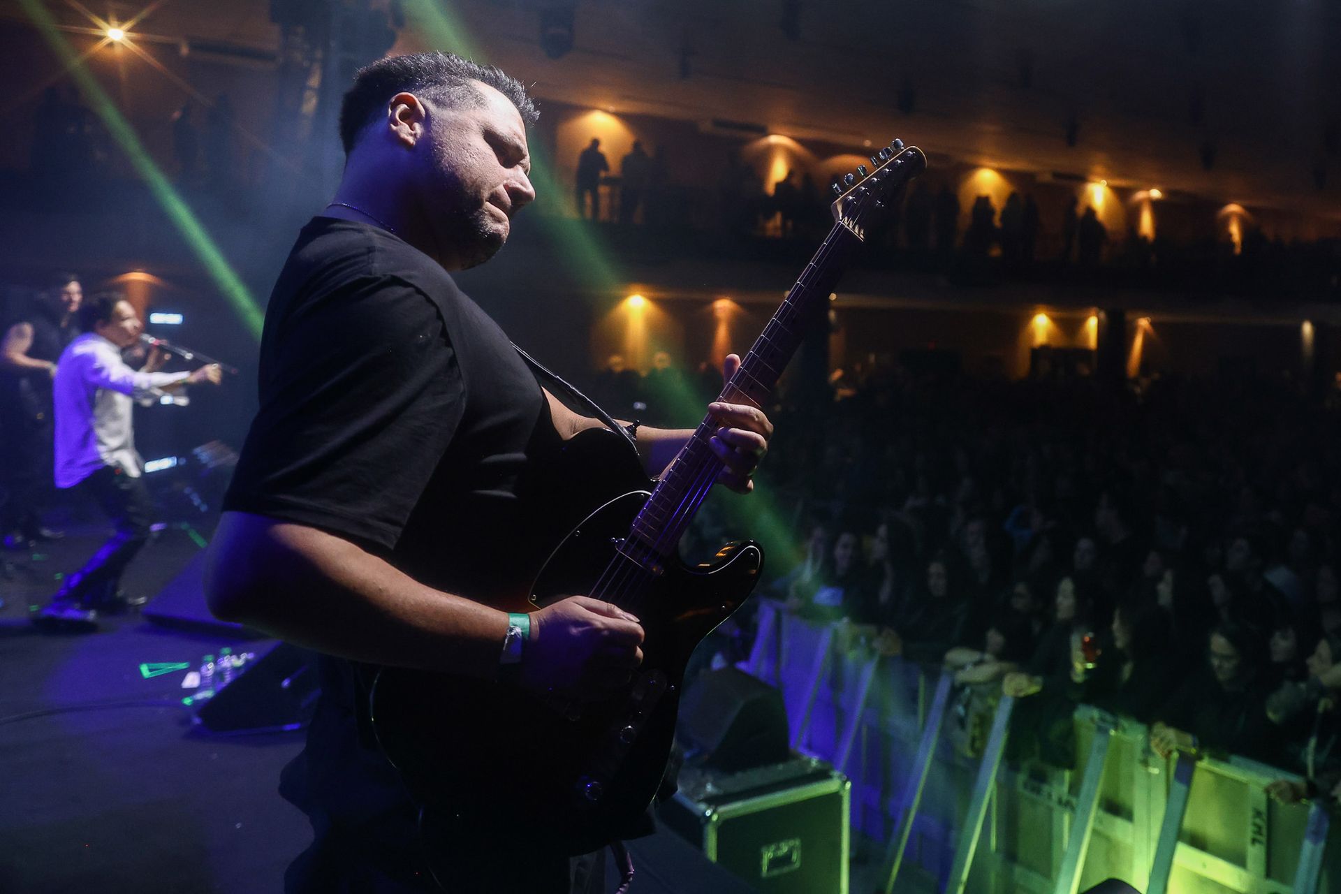 Um músico toca guitarra elétrica em um palco de concerto com pouca luz, enquanto outra pessoa se apresenta ao fundo.
