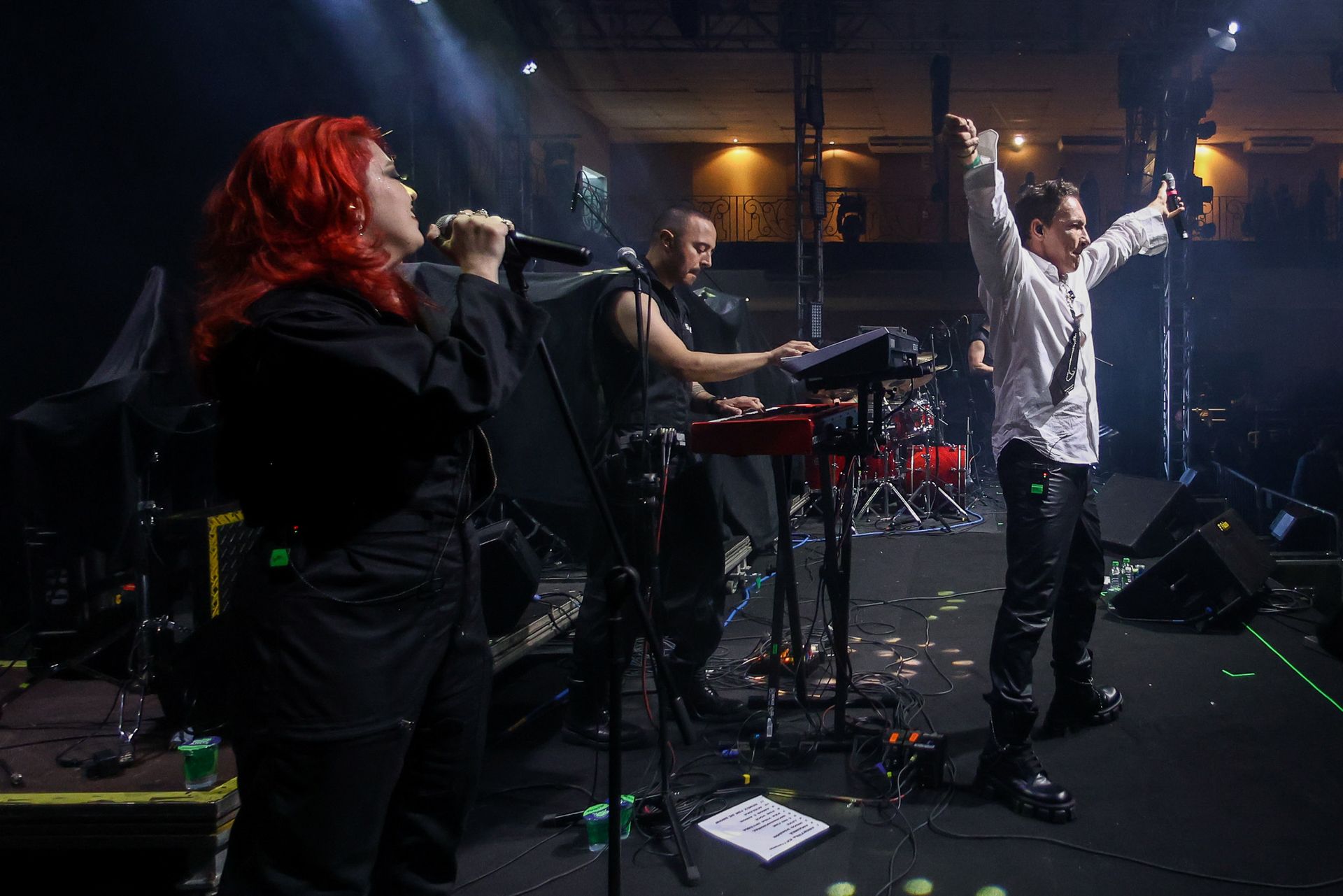 Uma banda se apresenta no palco: uma cantora ruiva com um microfone, um tecladista e uma pessoa com os braços erguidos.