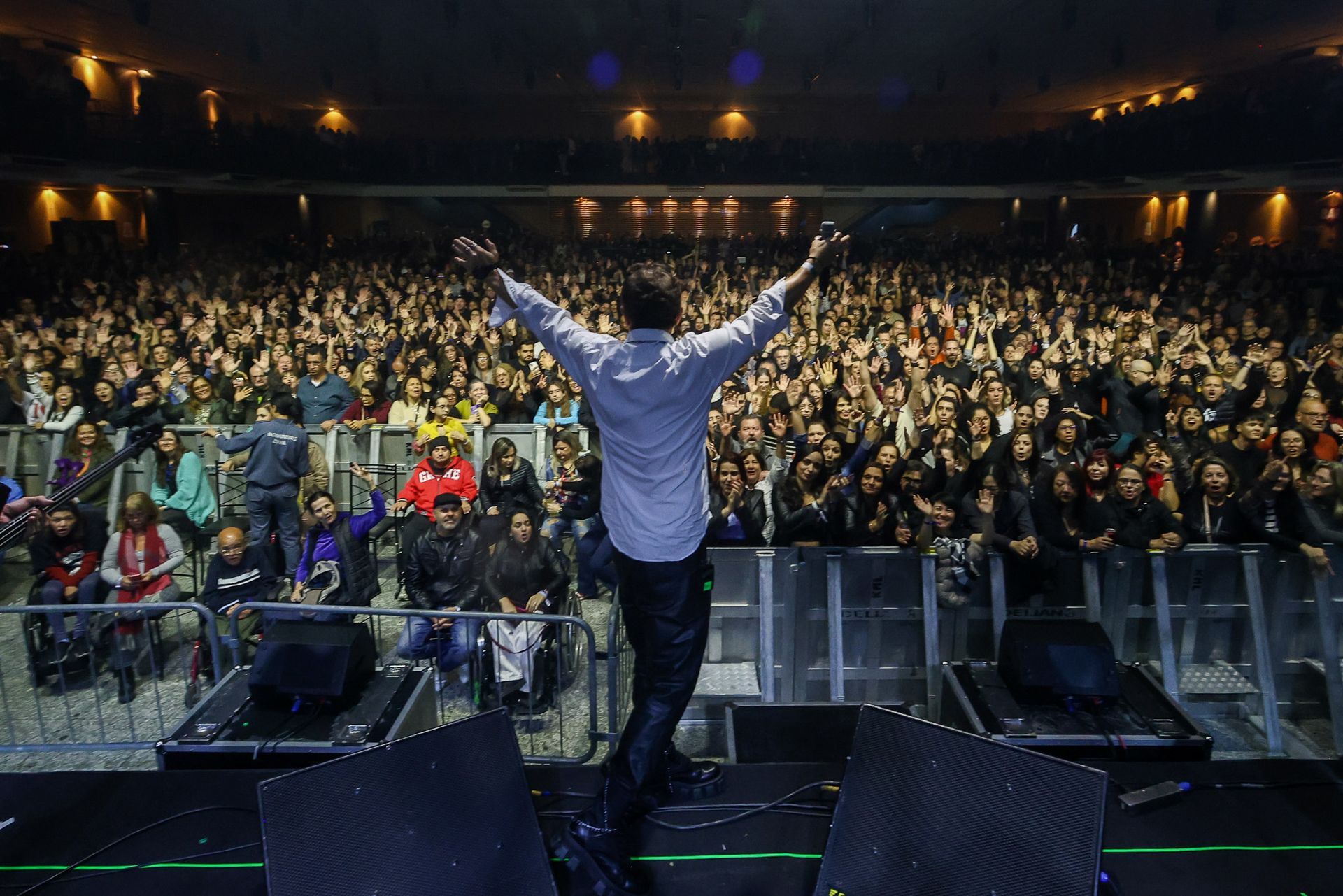 Um artista está em um palco com os braços estendidos, de frente para uma grande multidão que o aplaude.