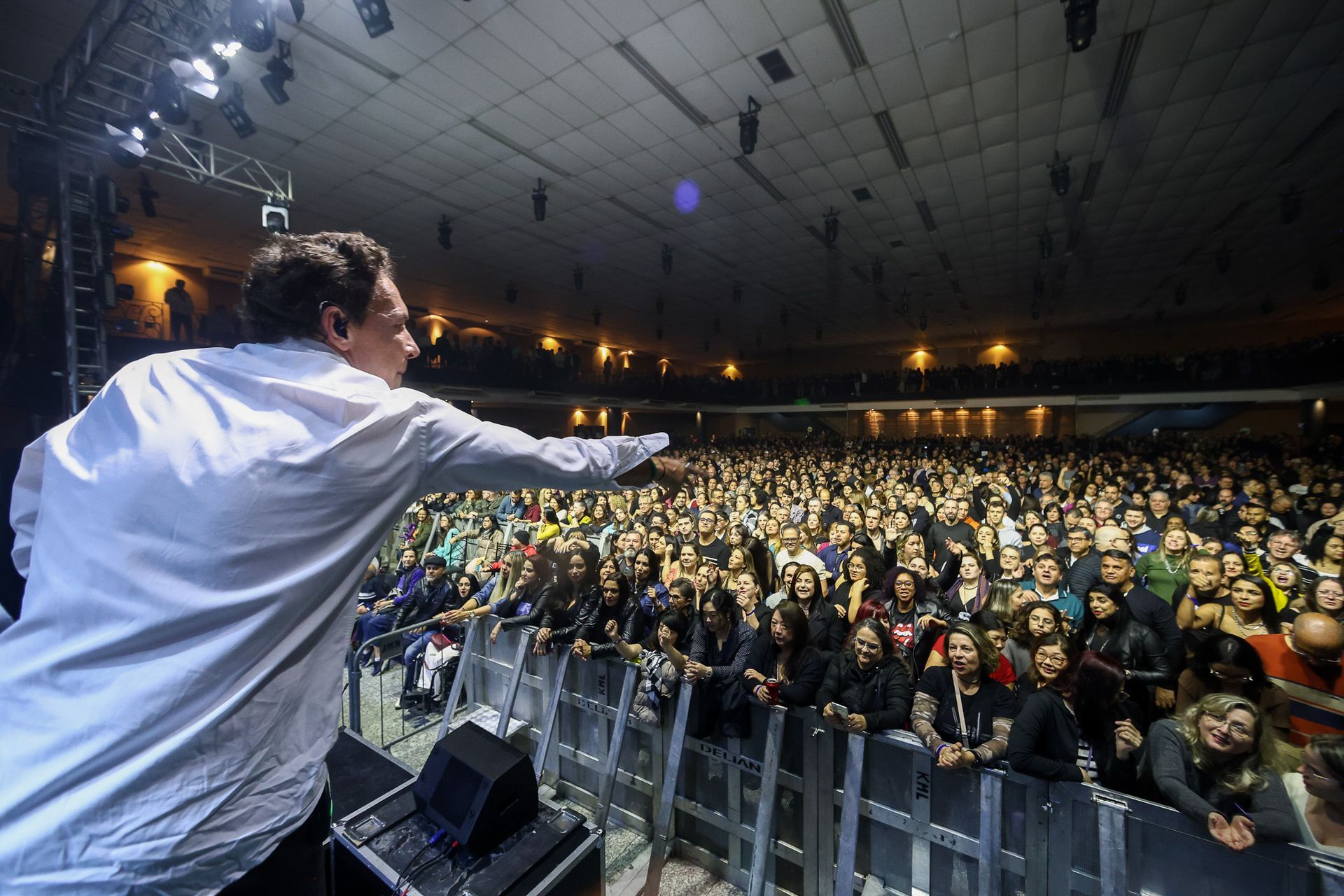Um artista de camisa branca aponta para uma grande multidão animada reunida em frente ao palco de uma sala de concertos.