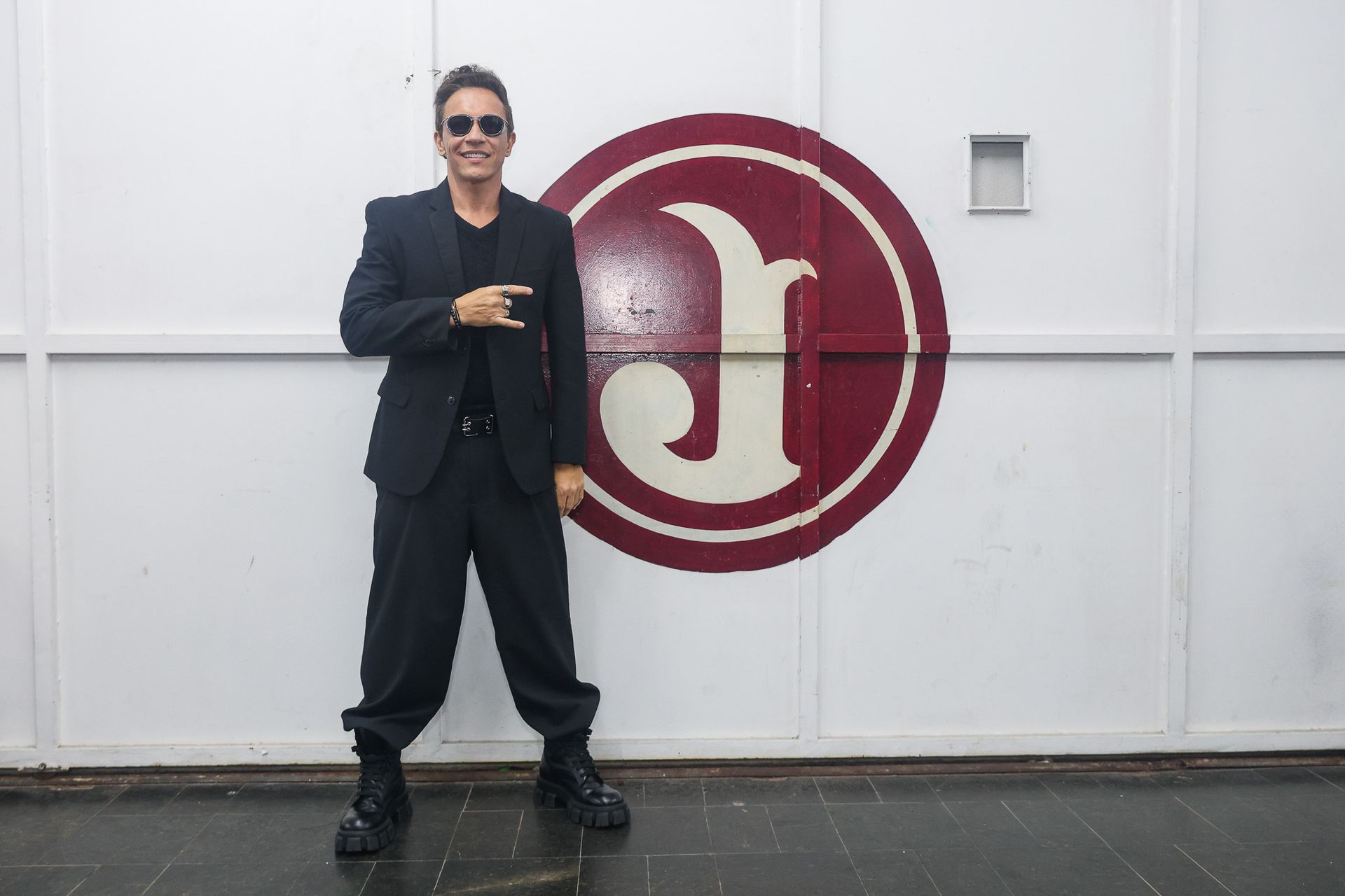 Um artista de terno preto e óculos escuros está em pé diante de uma parede branca com um grande logotipo circular vermelho.