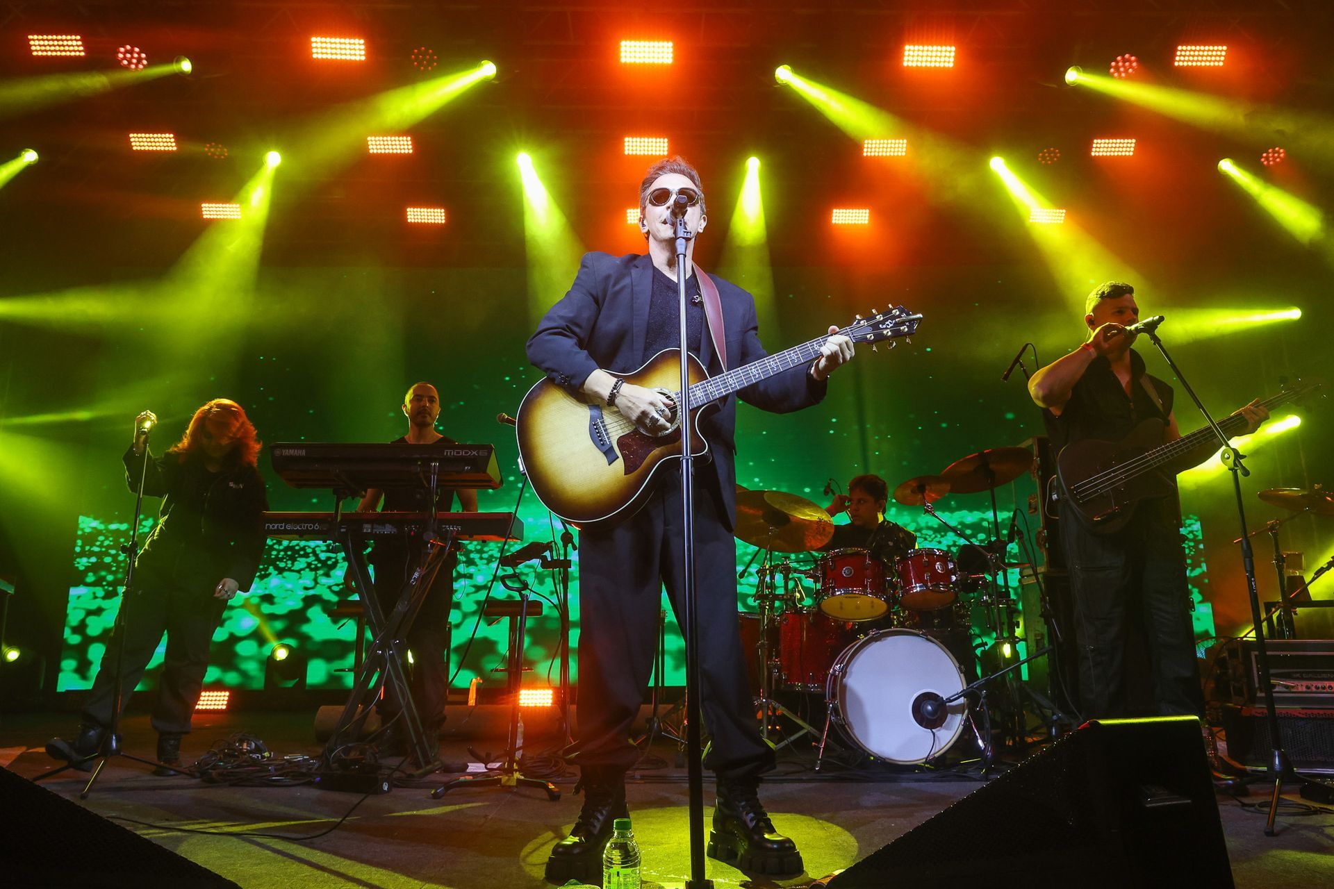 Uma banda de rock se apresenta em um palco banhado por luzes amarelas brilhantes, com o vocalista tocando violão.
