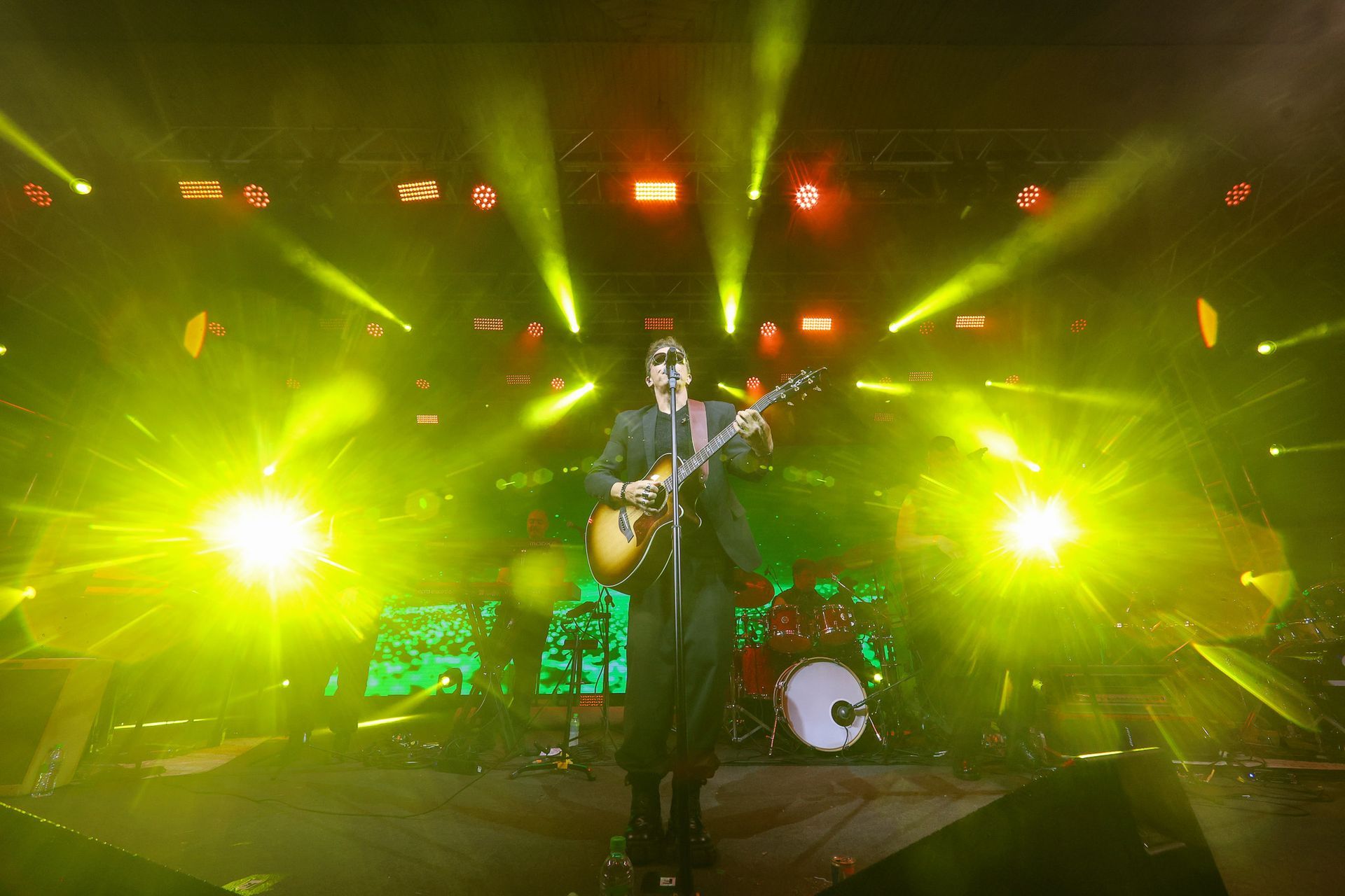 Um artista vestido de preto toca violão em um palco iluminado por holofotes amarelos brilhantes e névoa.