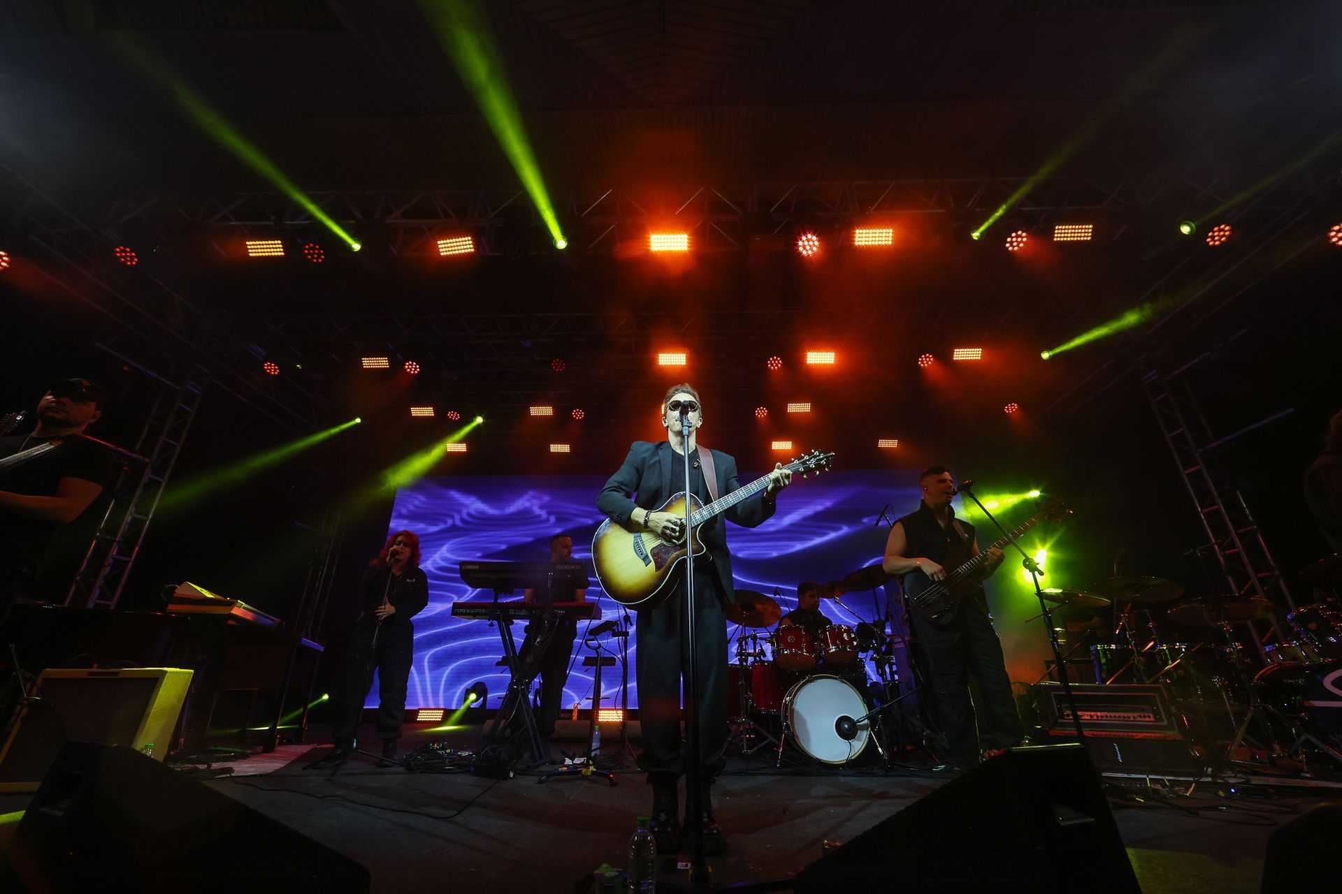Um artista cantando e tocando violão em um palco de concertos iluminado por luzes laranjas e feixes de luz verdes.