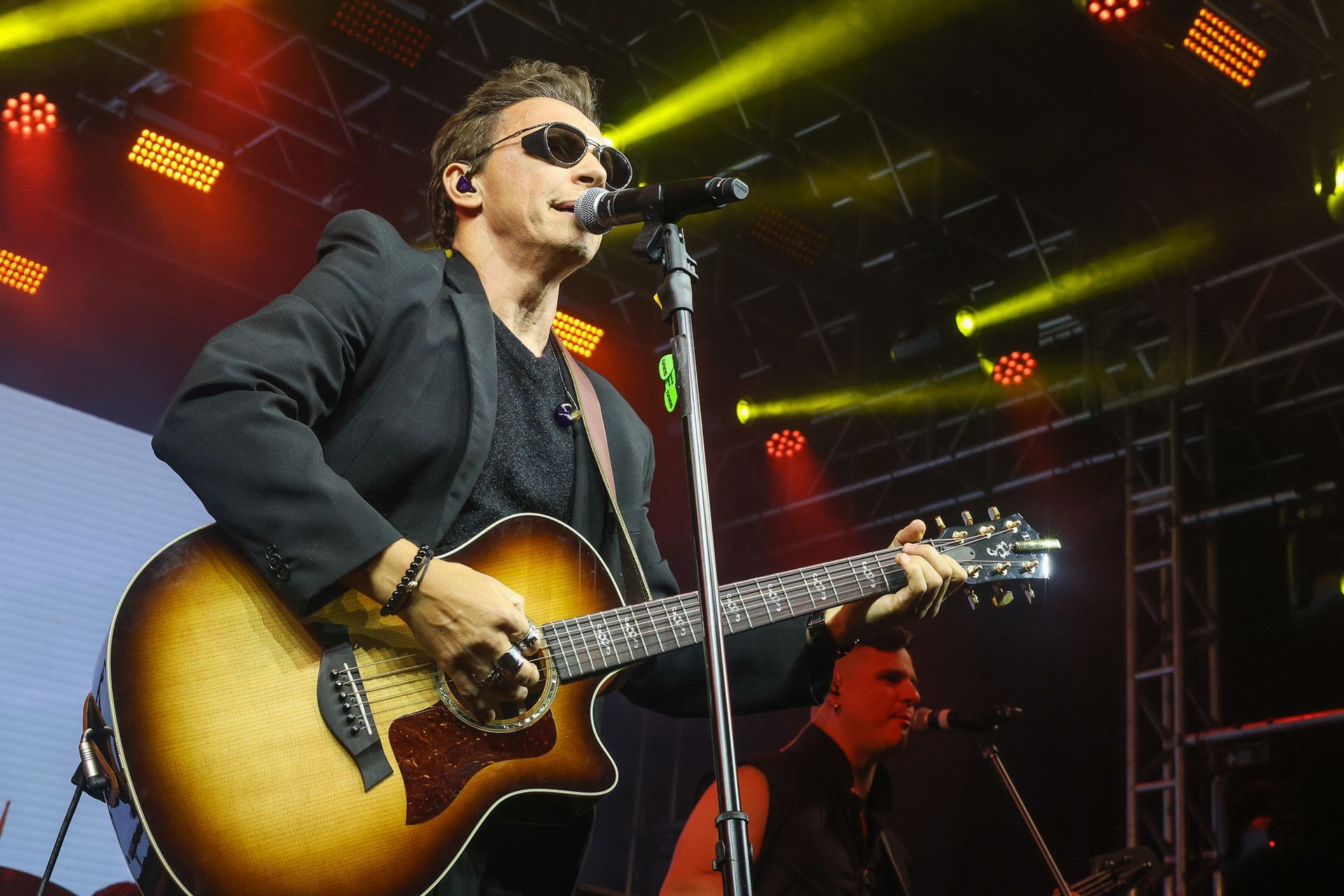 Um cantor de óculos escuros se apresenta no palco, tocando violão em um microfone sob luzes coloridas de show.