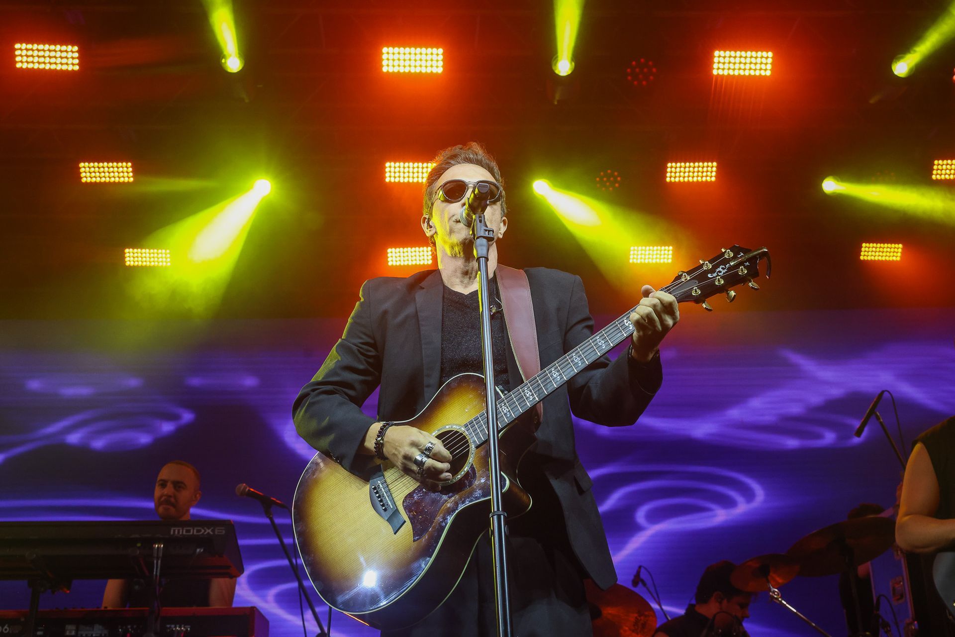 Um músico de jaqueta preta e óculos escuros tocando violão em um palco com luzes amarelas brilhantes.