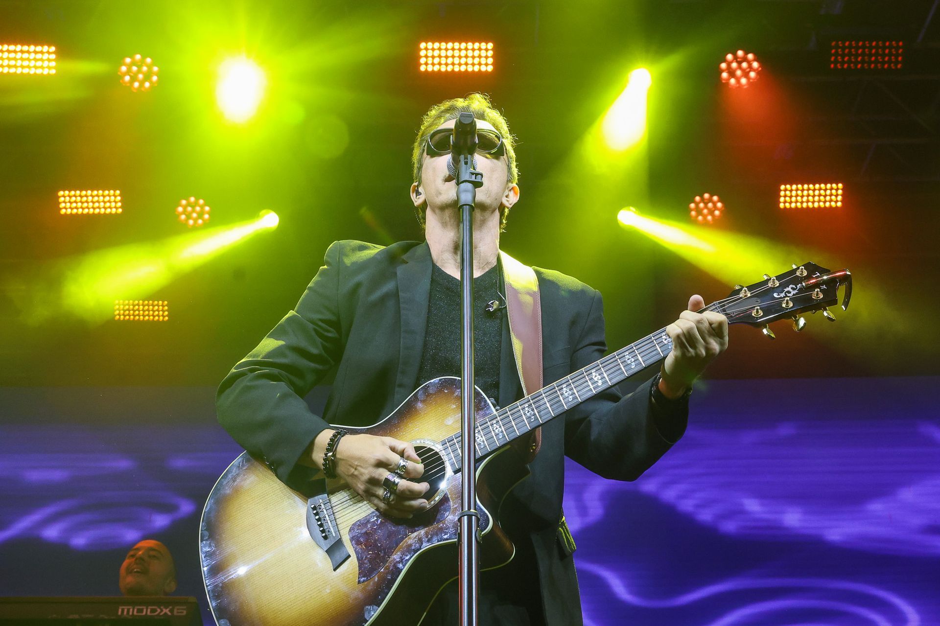 Um artista de terno preto e óculos escuros toca violão enquanto canta ao microfone no palco.