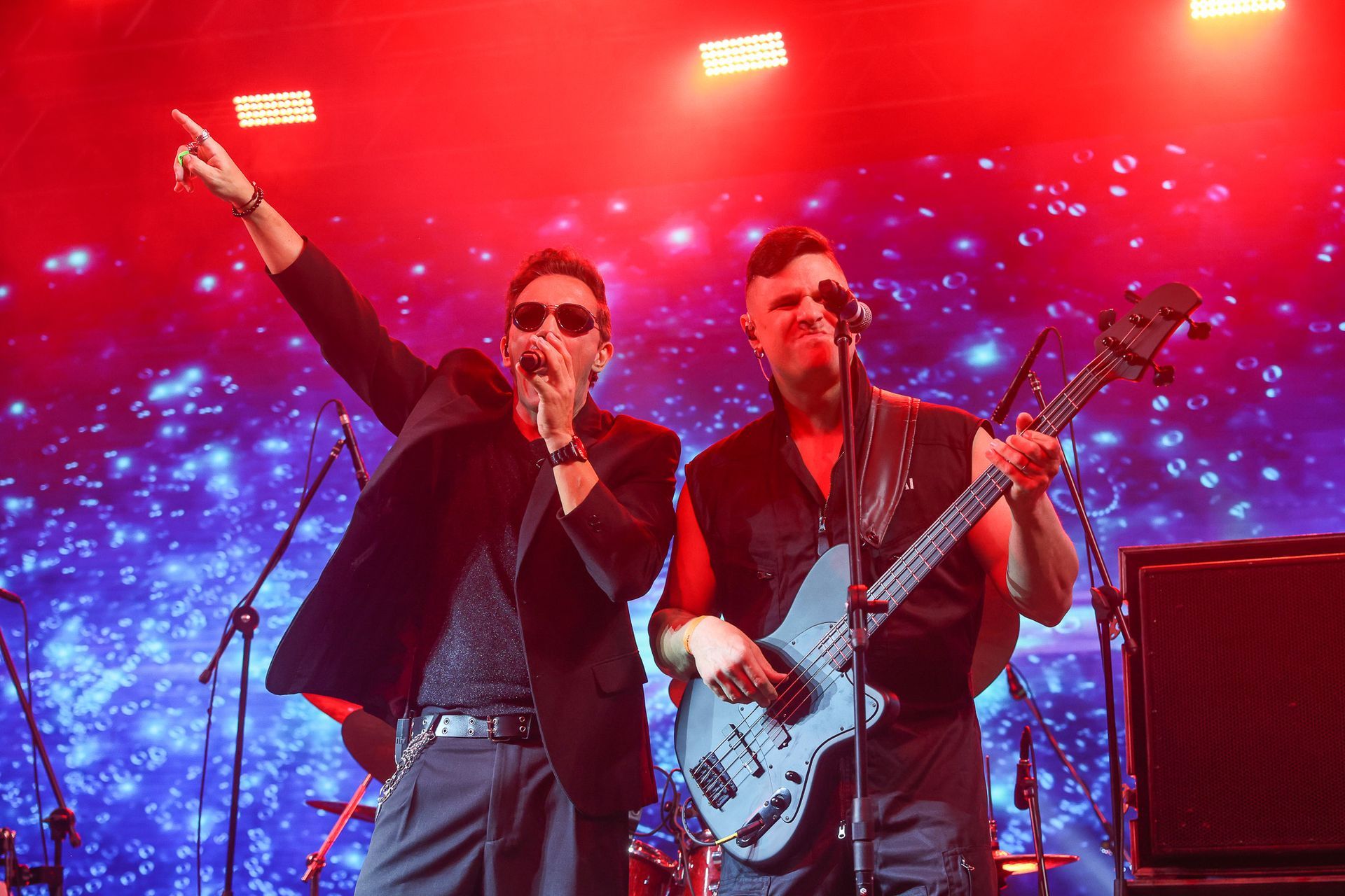 Dois músicos se apresentam no palco sob uma iluminação vermelha intensa; um canta ao microfone, o outro toca um baixo azul.