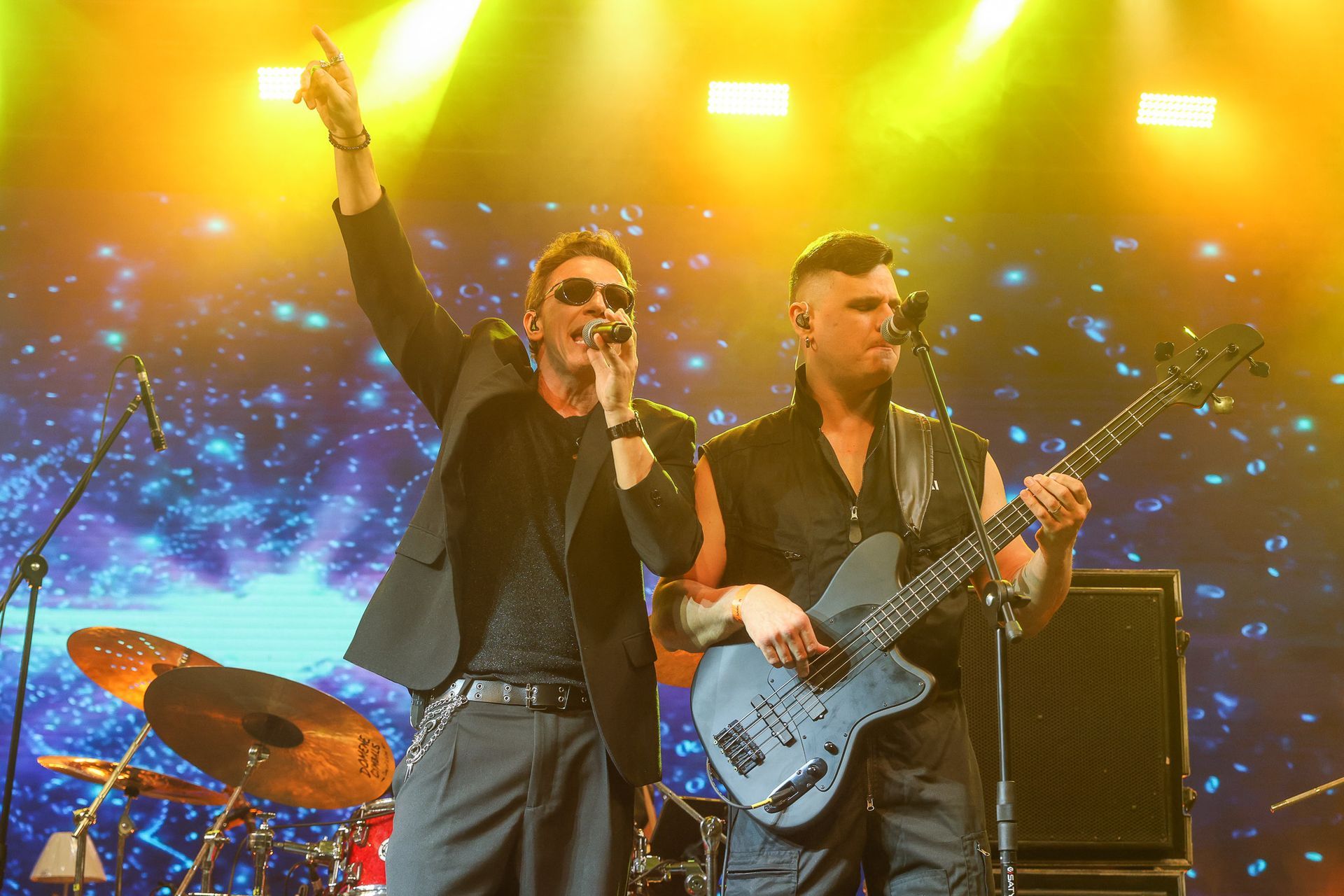 Dois músicos se apresentam no palco contra um cenário brilhante e estrelado; um canta o outro toca baixo.