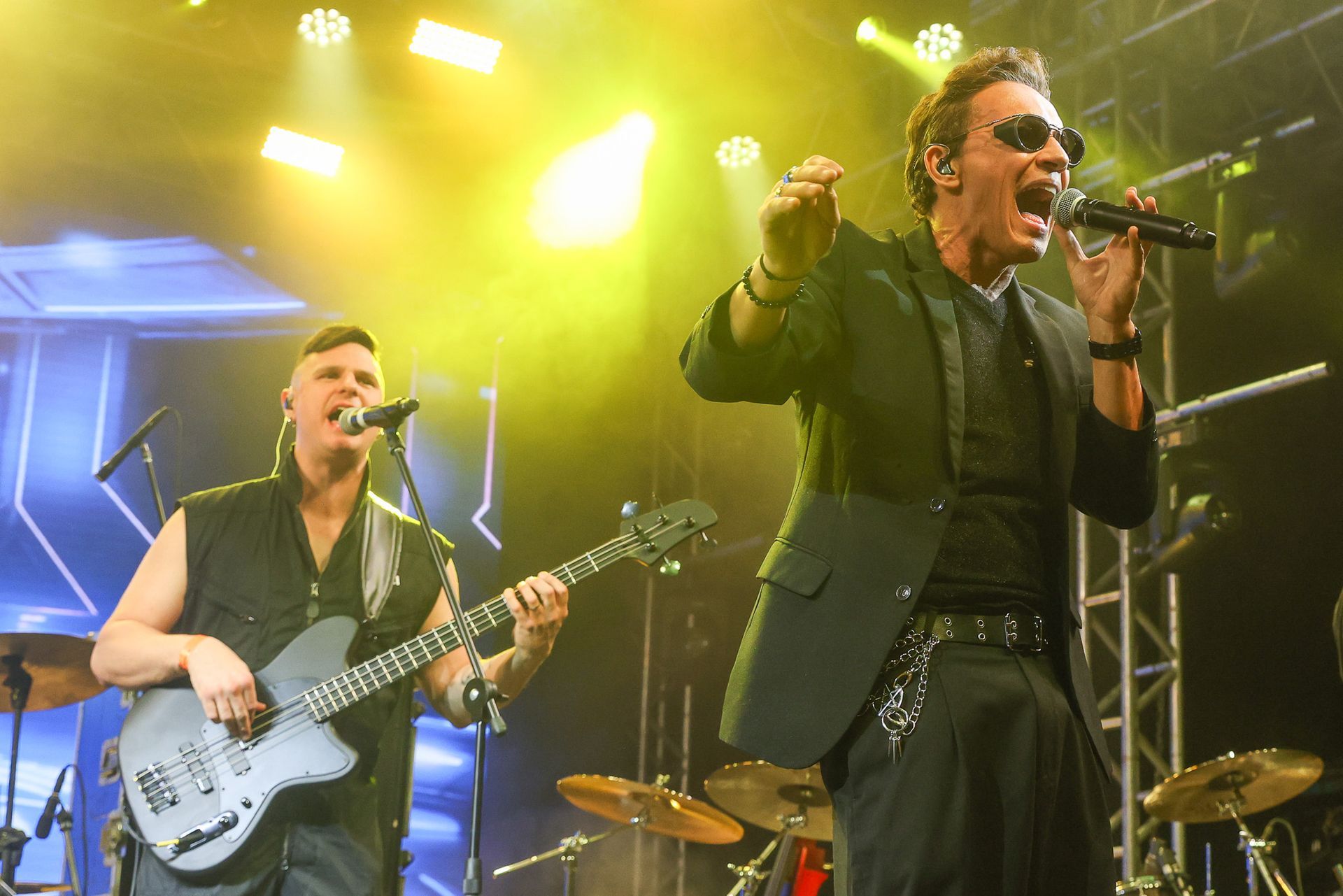 Dois integrantes da banda se apresentam em um palco bem iluminado; um baixista e o vocalista.