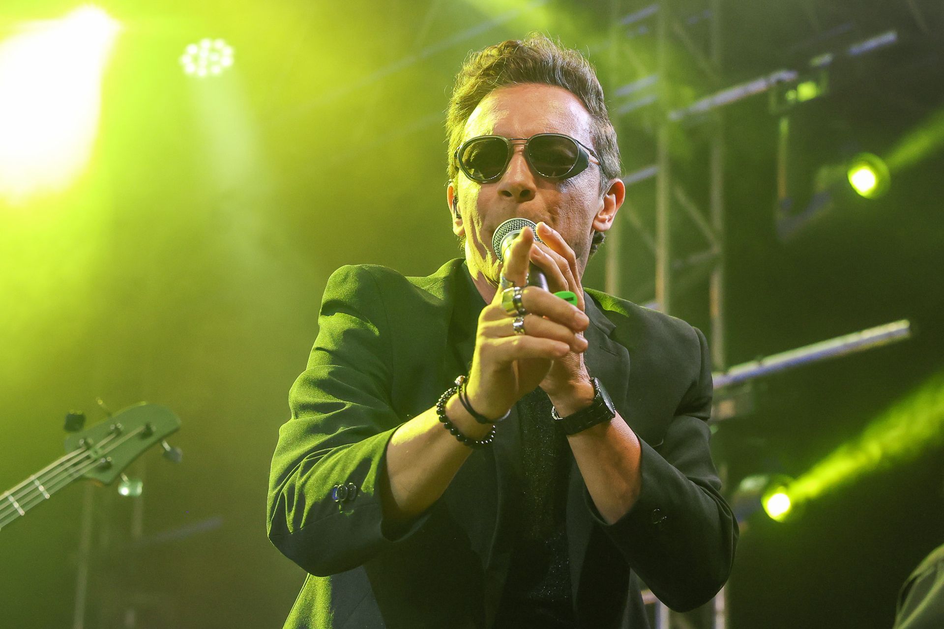 Um artista usando óculos escuros canta em um microfone no palco sob holofotes amarelos brilhantes.
