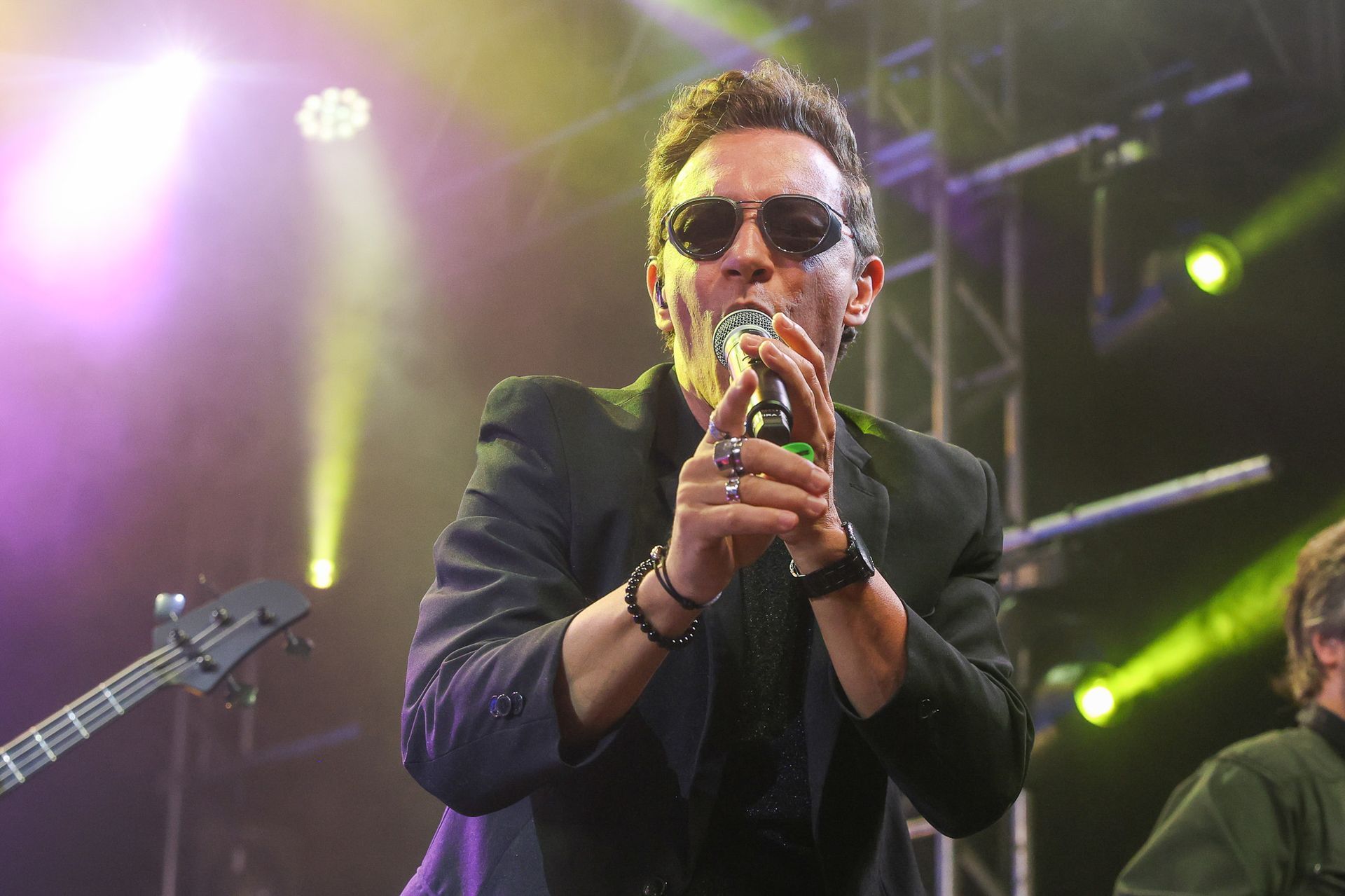 Um músico usando óculos escuros canta em um microfone no palco sob luzes de palco brilhantes e coloridas.