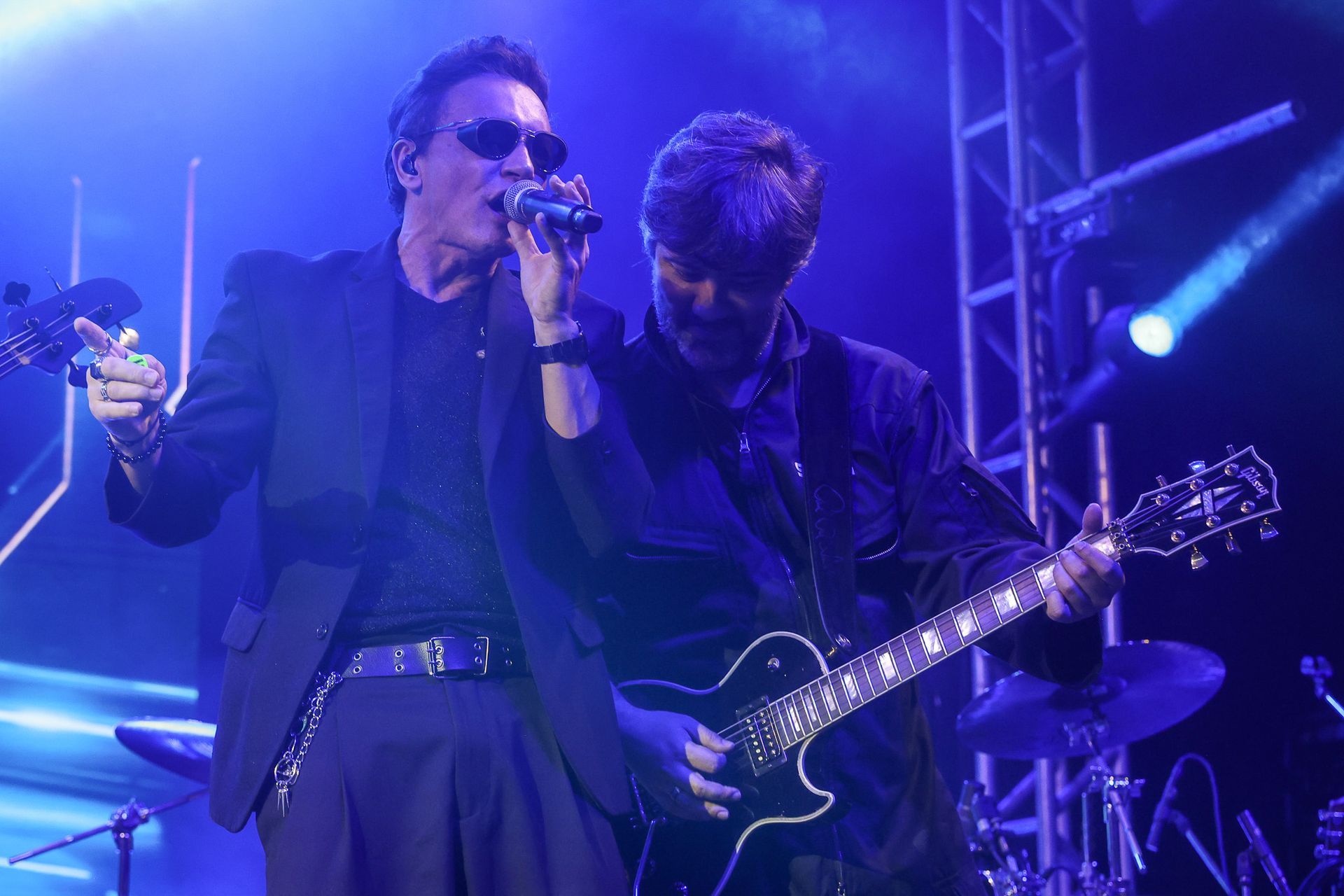 Dois músicos se apresentam no palco banhados por luz azul: um canta ao microfone enquanto o outro toca guitarra elétrica.