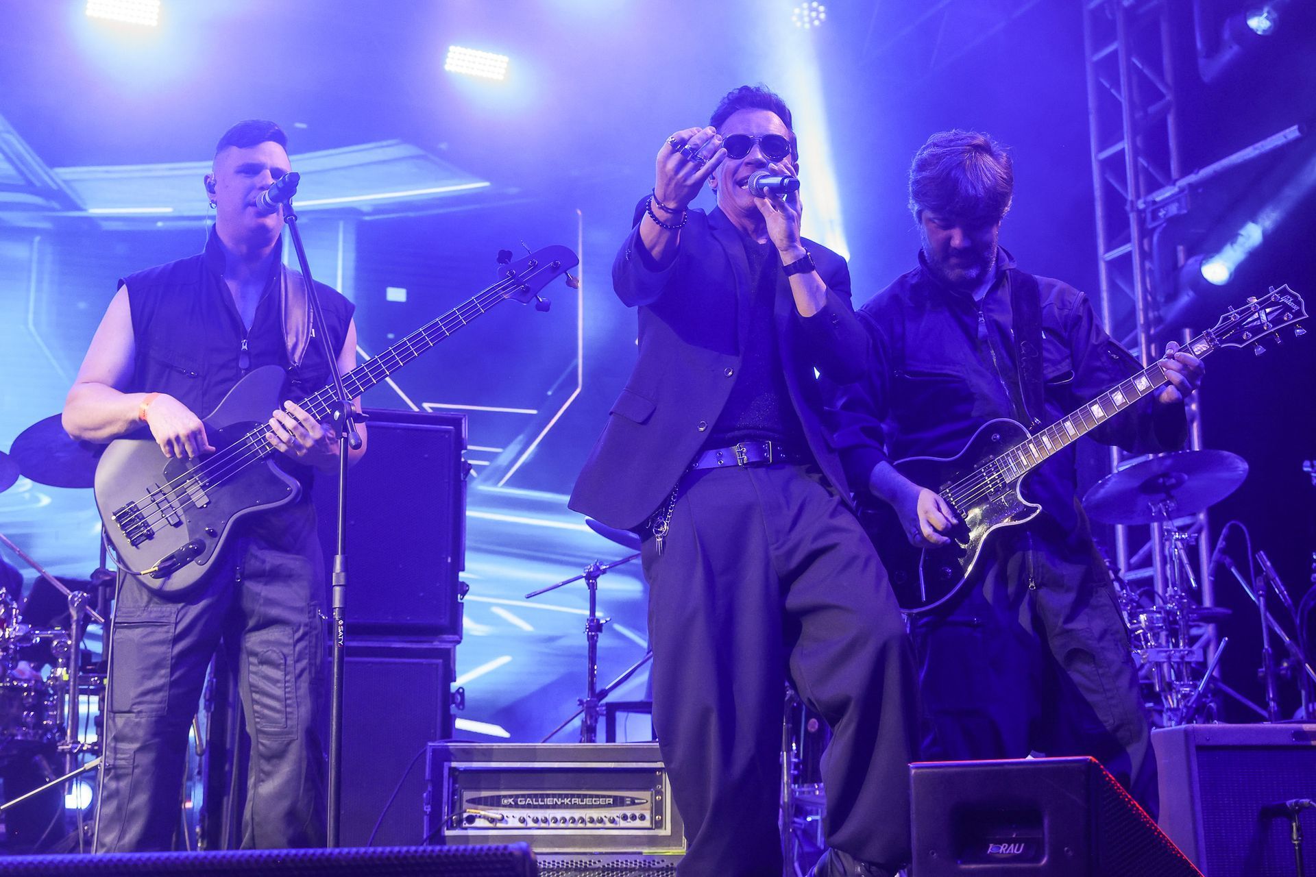 Uma banda se apresenta no palco sob luzes roxas, com dois guitarristas e um vocalista segurando um microfone.