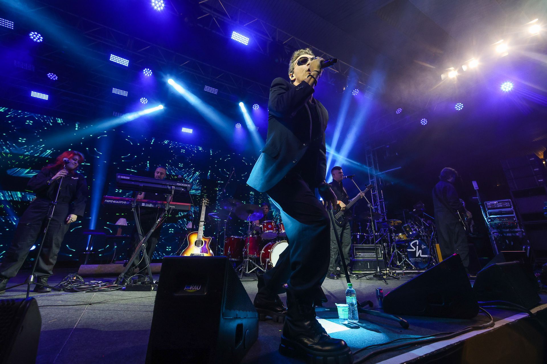 Um cantor se apresenta no palco durante um show, banhada por holofotes azuis, com uma banda tocando instrumentos ao fundo.