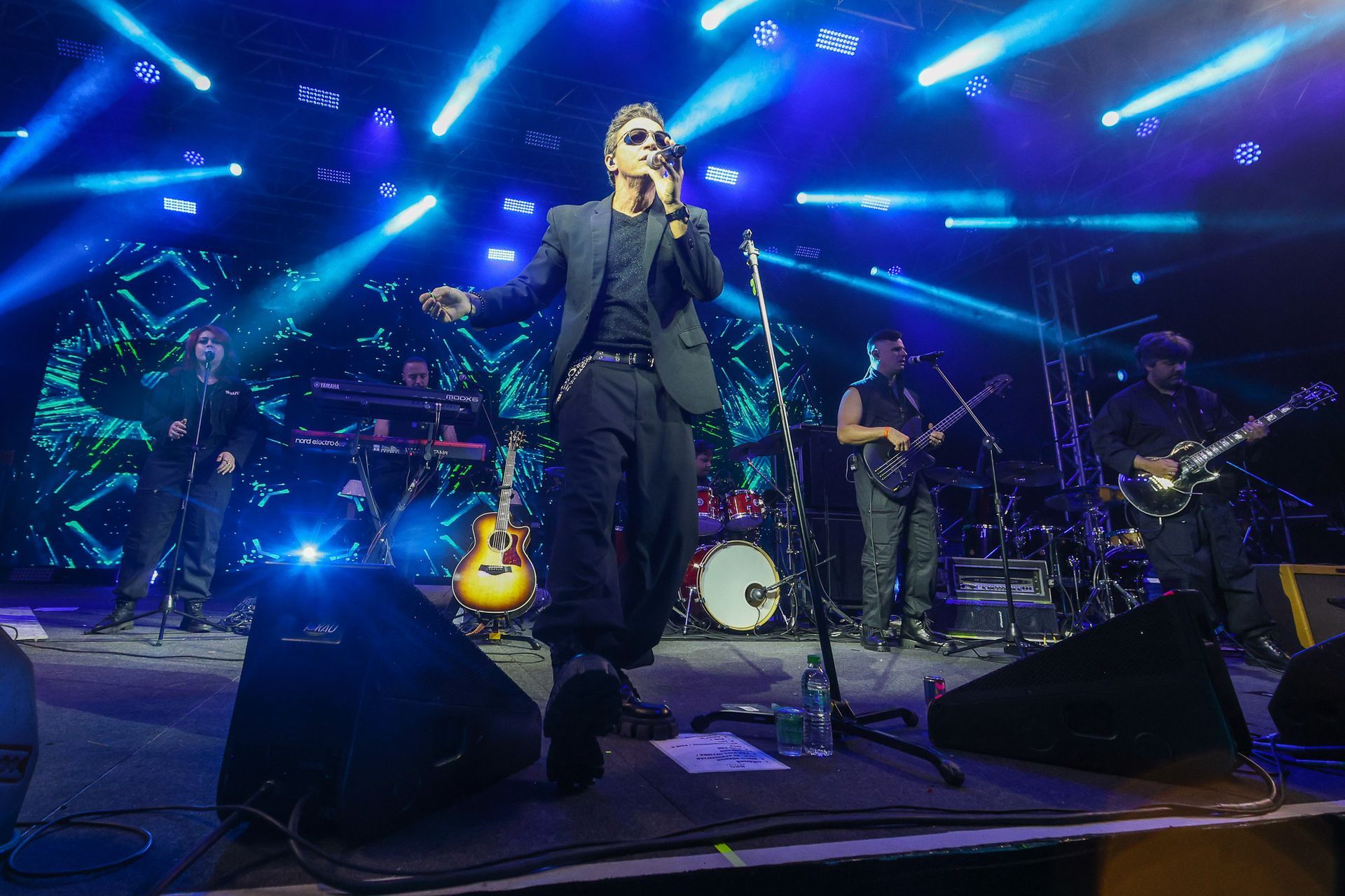 O vocalista, de blazer e óculos escuros, se apresenta no palco com uma banda sob holofotes azuis brilhantes e um telão.