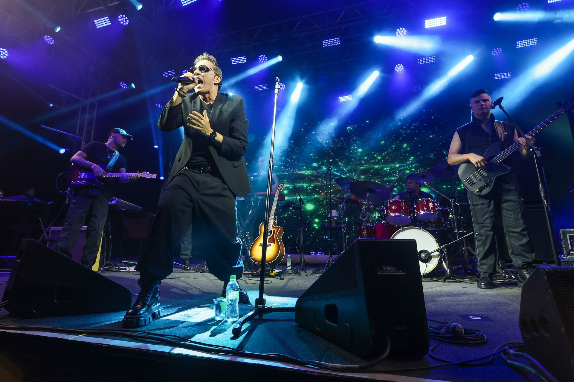 Uma banda de rock se apresenta em um palco sob holofotes azuis brilhantes, com o vocalista gesticulando enquanto canta.