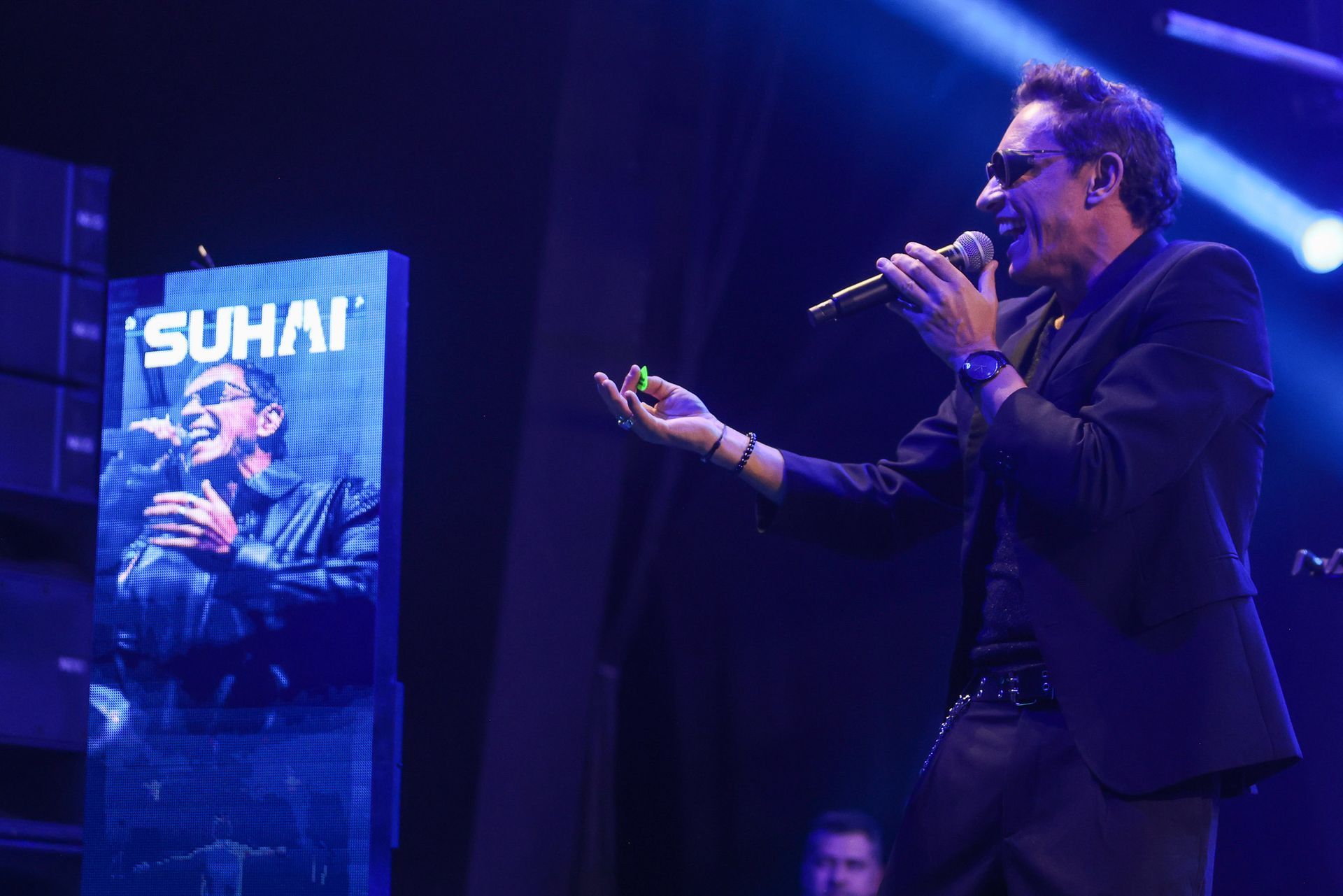 Um homem de terno escuro canta no palco, ao lado de uma grande tela vertical que exibe sua imagem e a palavra 