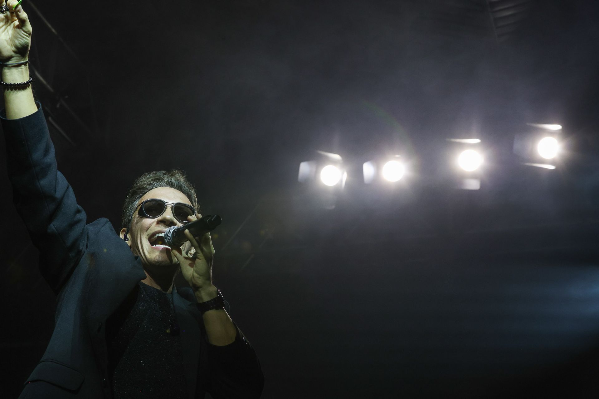 Um artista de terno escuro e óculos escuros cantando em um microfone no palco, iluminada por holofotes brilhantes.