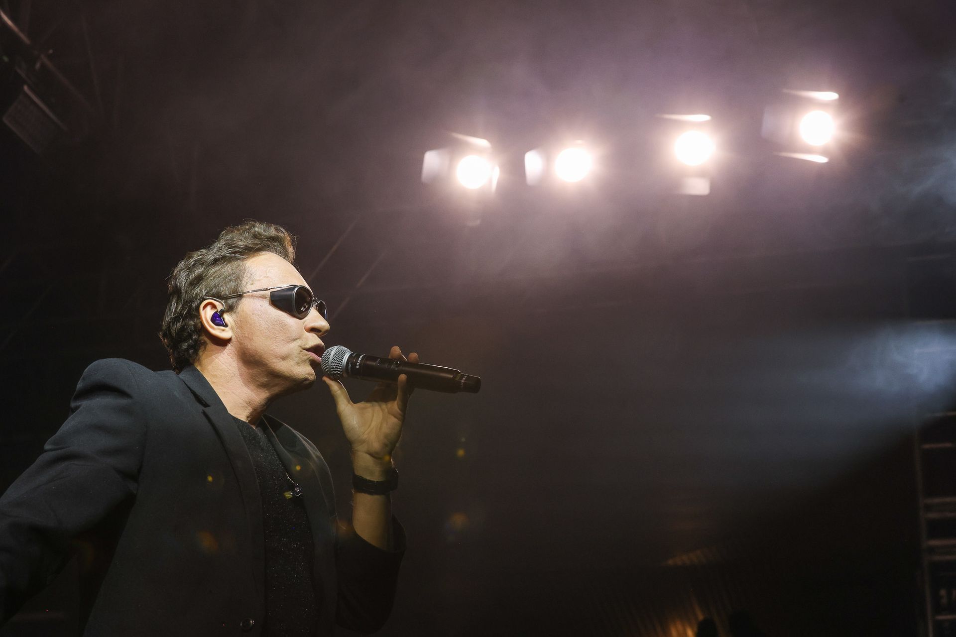 Um cantor de blazer escuro e óculos escuros se apresenta no palco, falando ao microfone sob luzes de palco brilhantes.