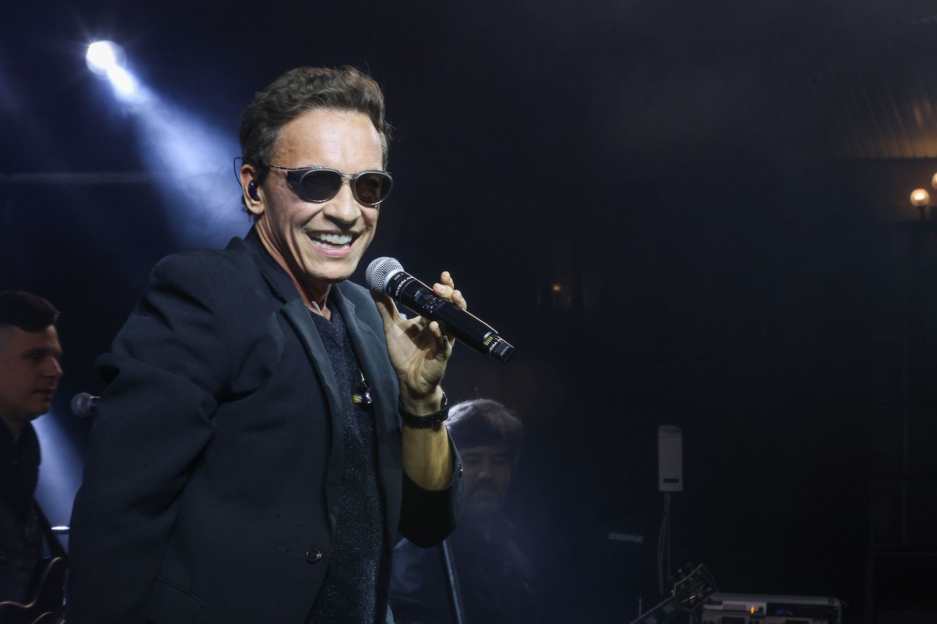 Um artista de terno preto e óculos escuros sorri enquanto canta ao microfone em um palco sob um holofote.