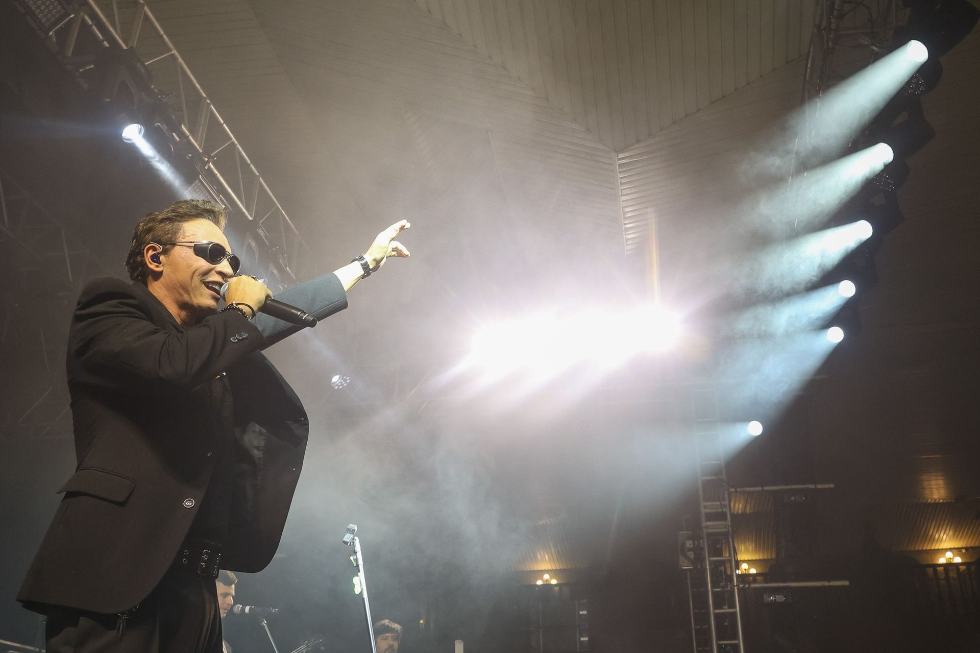Um cantor de terno e óculos escuros se apresenta em um palco, gesticulando em direção aos holofotes brilhantes.