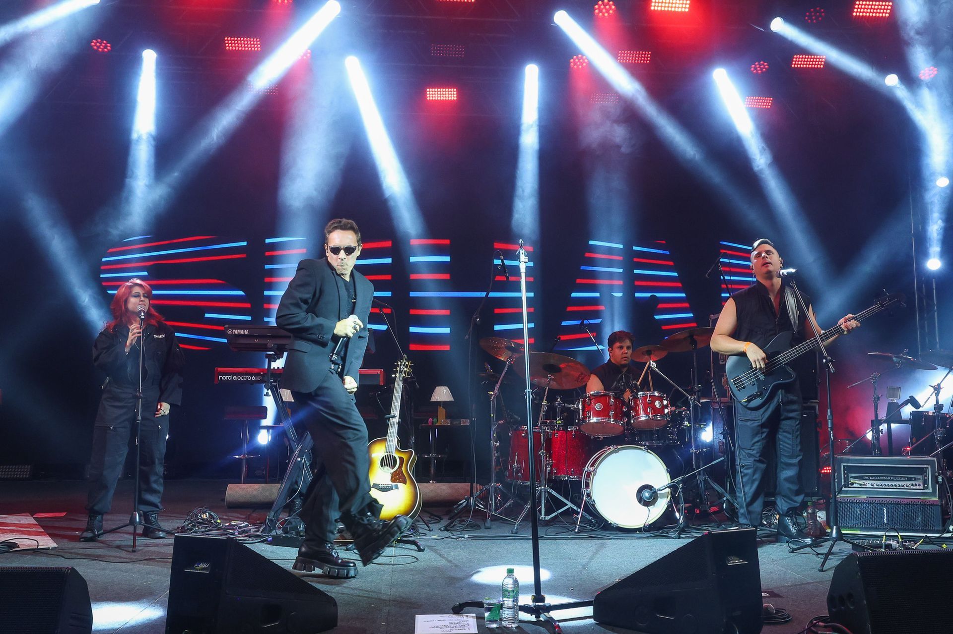 Uma banda se apresenta em um palco, composta por um vocalista de terno, um baterista e um guitarrista.