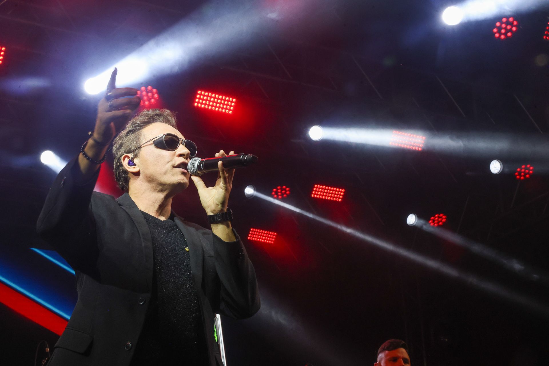 Um artista de terno preto e óculos escuros canta em um microfone no palco, iluminado por holofotes brilhantes.