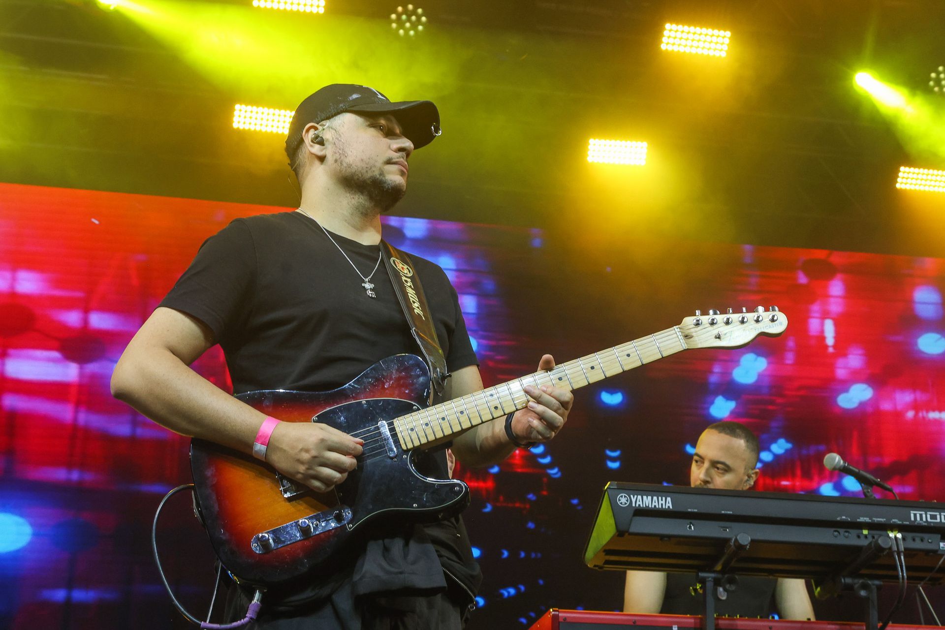 Um guitarrista toca uma guitarra em um palco com iluminação brilhante e colorida, enquanto um tecladista toca próximo a ele.