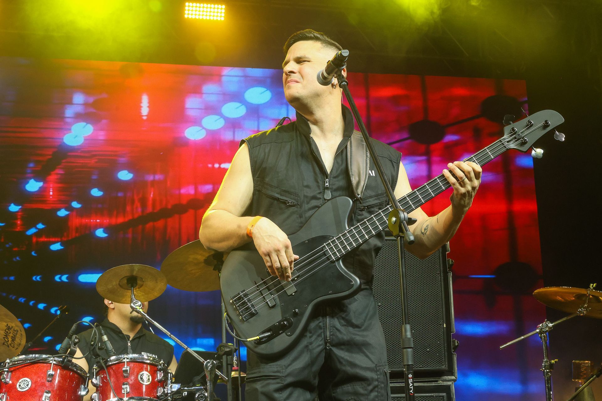 Um músico tocando um baixo preto no palco em frente a uma tela vermelha e azul, com um baterista visível nas proximidades.