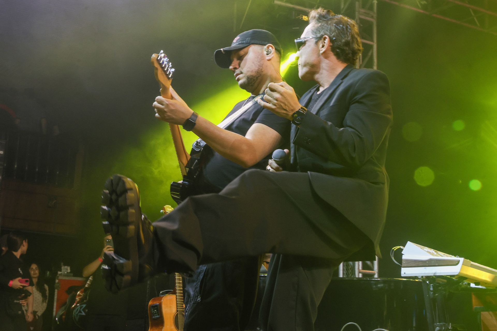 Dois músicos se apresentam no palco; um toca guitarra elétrica enquanto o outro canta, ambos capturados em uma pose enérgica.