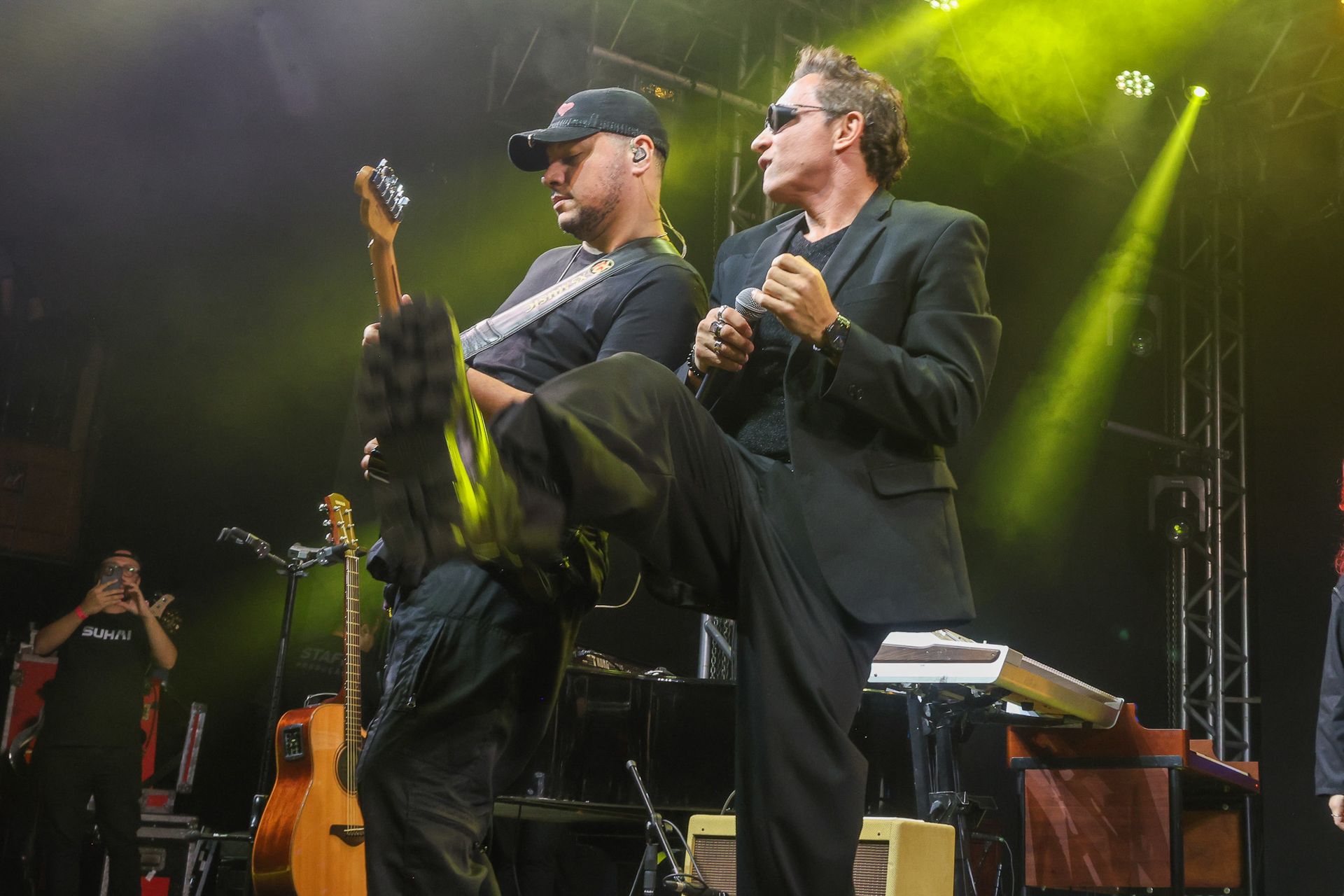 Um músico toca guitarra enquanto uma vocalista de terno preto se apresenta com ele em um palco de concerto bem iluminado.