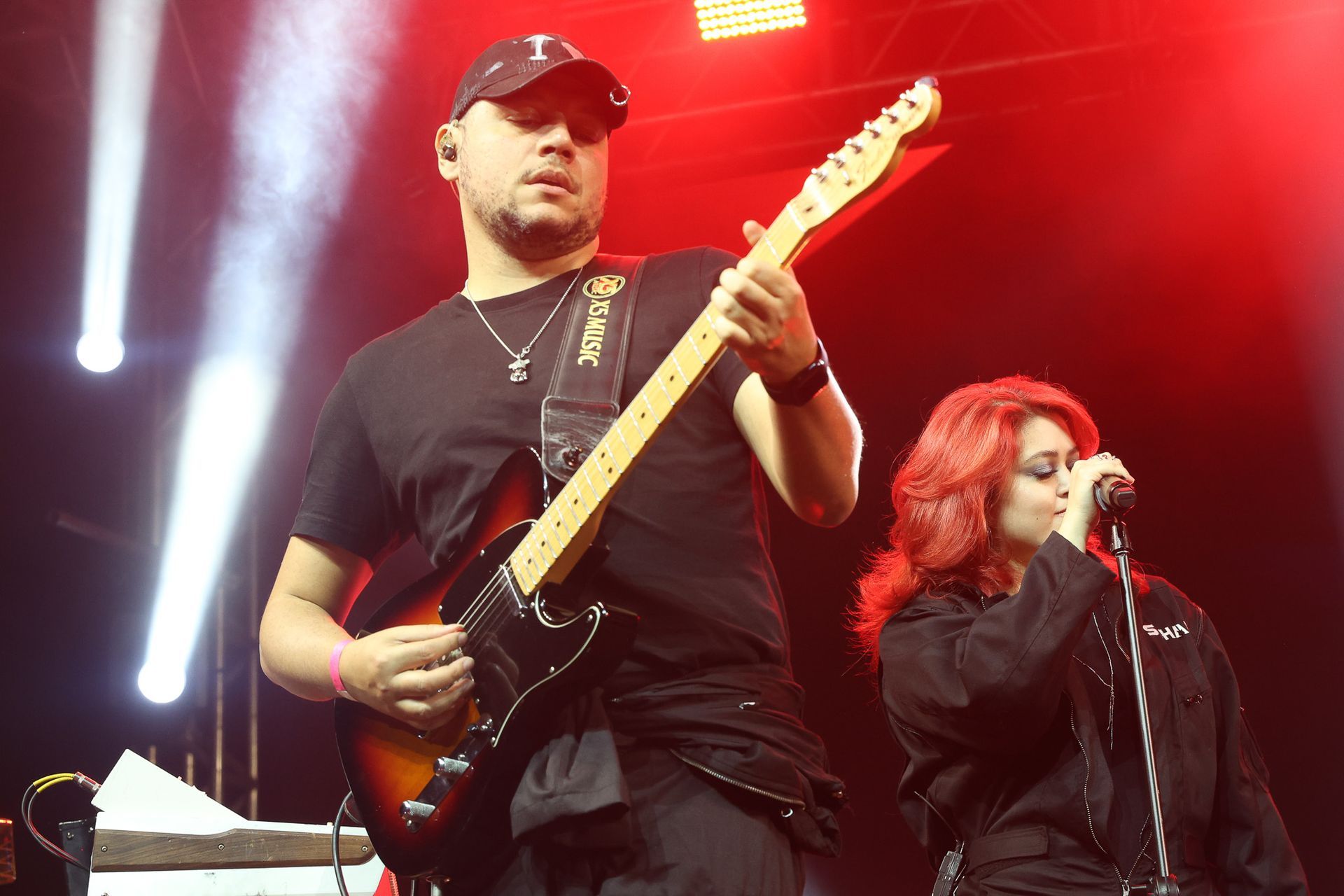 Um músico toca guitarra elétrica ao lado de uma cantora que segura um microfone em um palco com iluminação vermelha.