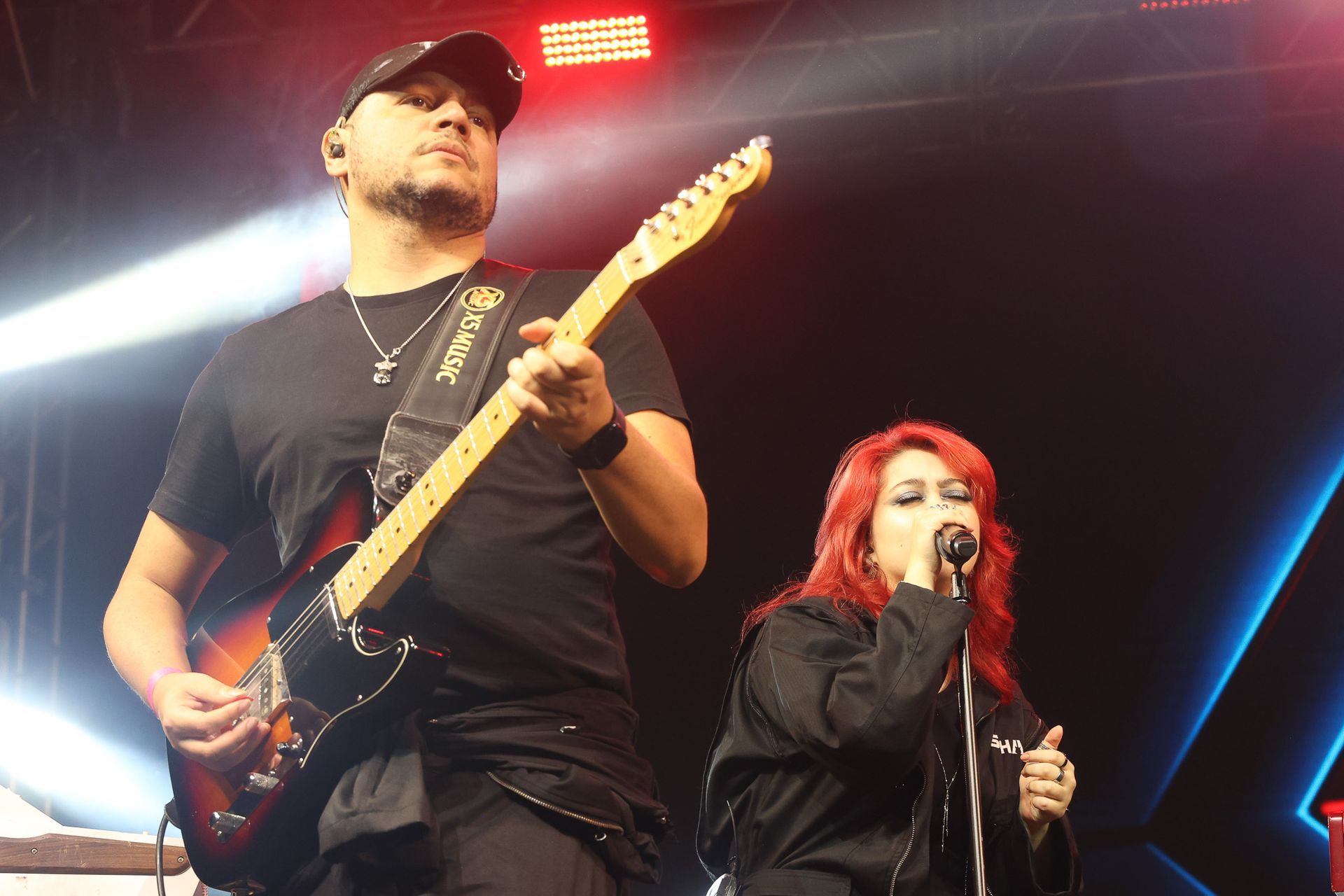 Um guitarrista usando boné e camiseta preta se apresenta ao lado de uma cantora ruiva de jaqueta preta em um palco.