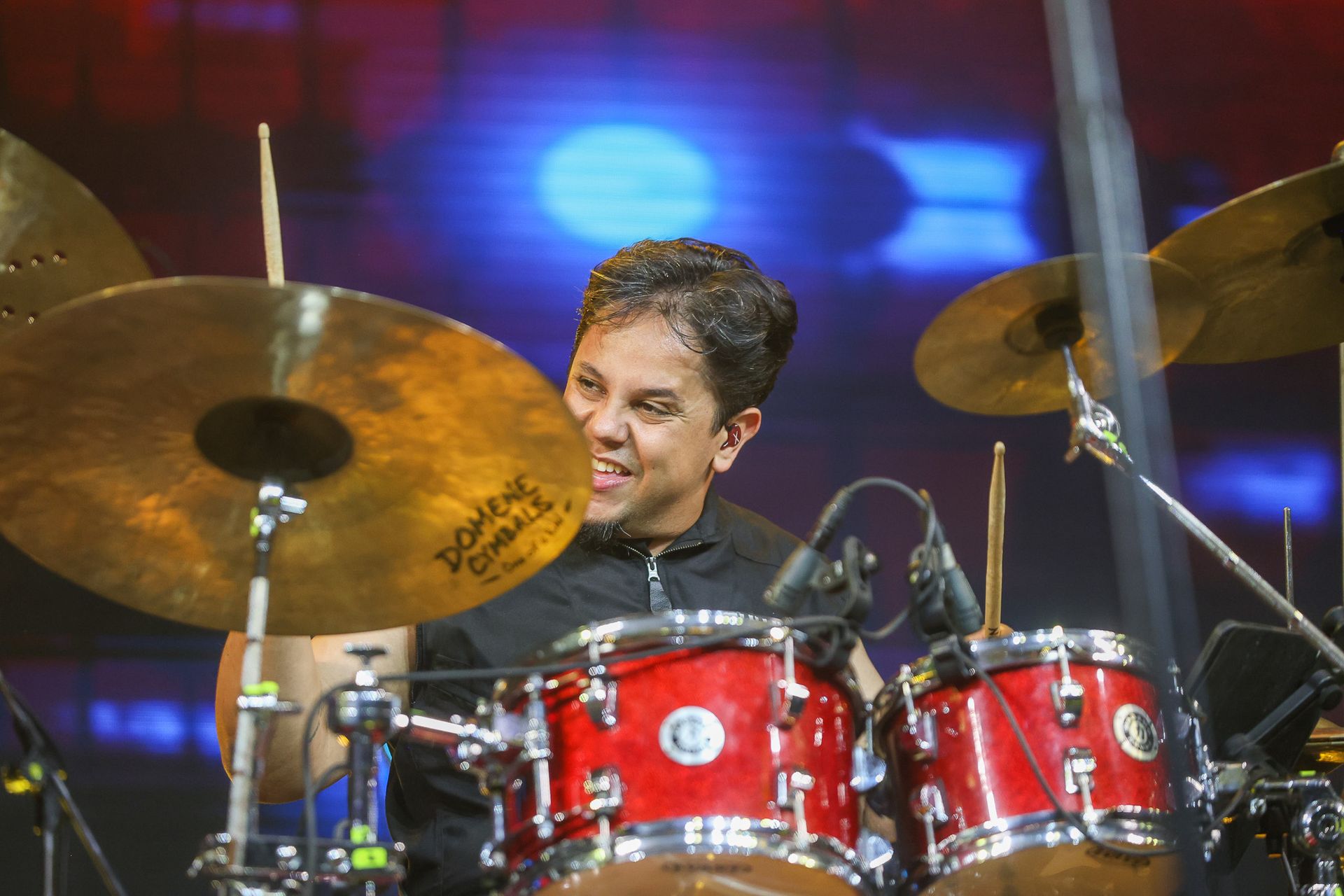 Um baterista sorridente tocando bateria e pratos vermelhos em um palco com luzes azuis e vermelhas desfocadas ao fundo.