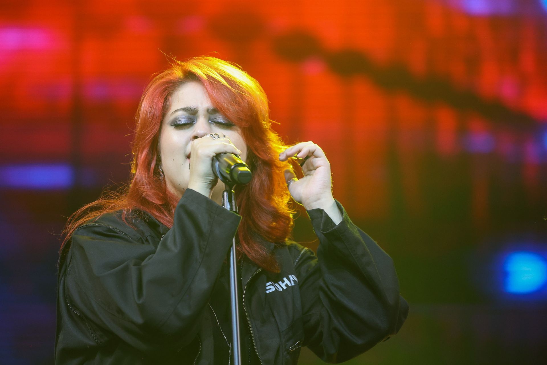 Uma cantora ruiva, de olhos fechados, canta em um microfone contra um vibrante fundo de palco em tons de laranja.