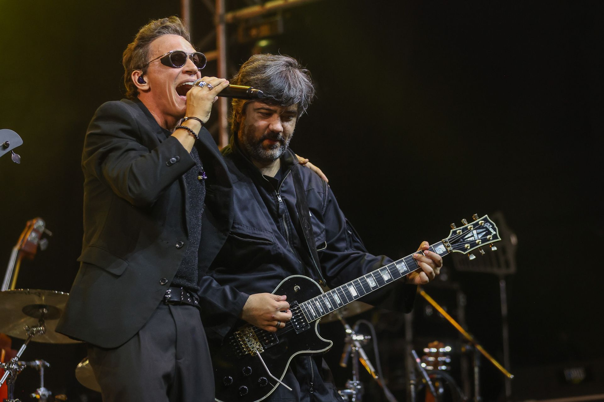 Dois músicos se apresentando no palco: um canta ao microfone enquanto o outro toca guitarra elétrica.