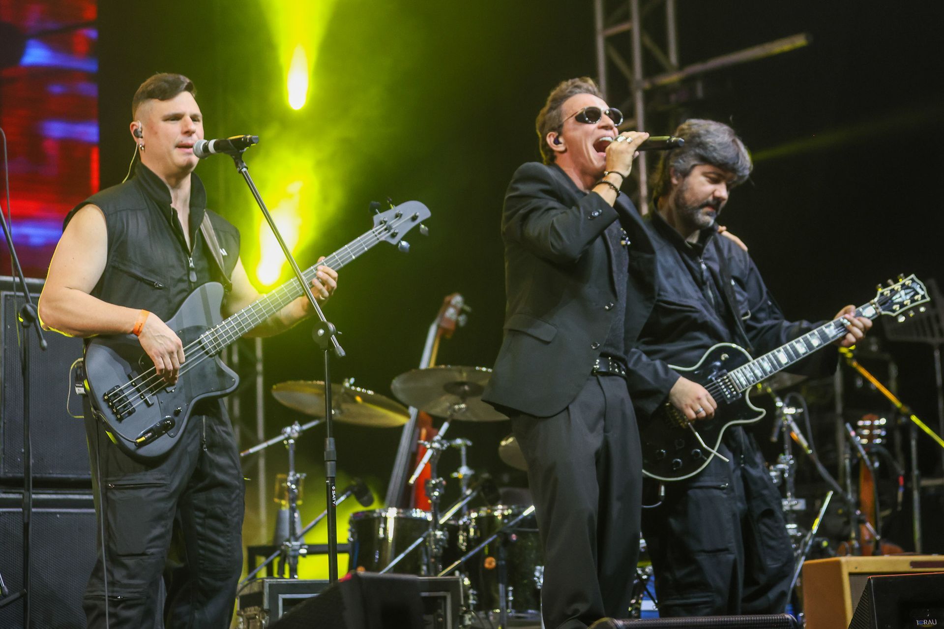 Um trio musical se apresenta em um palco com pouca luz, cantando e tocando guitarra elétrica e baixo sob luzes brilhantes.