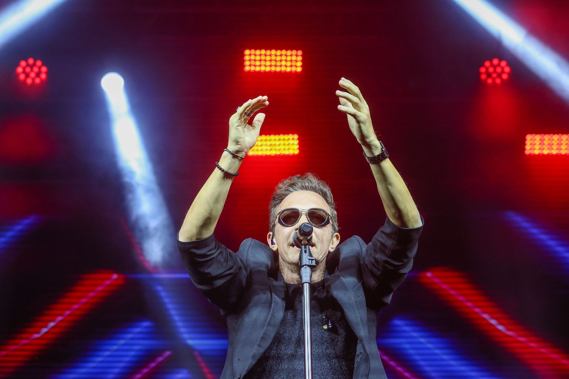 Cantor de óculos escuros, com os braços erguidos, cantando ao microfone em um palco com luzes vermelhas e azuis brilhantes.