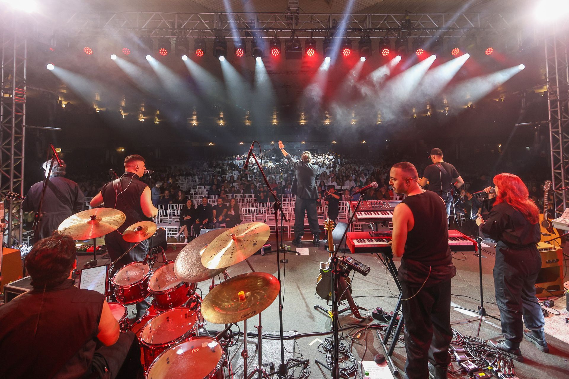 Uma banda se apresenta em um show com luzes brilhantes, bateria e instrumentos diante de uma grande plateia com iluminação.
