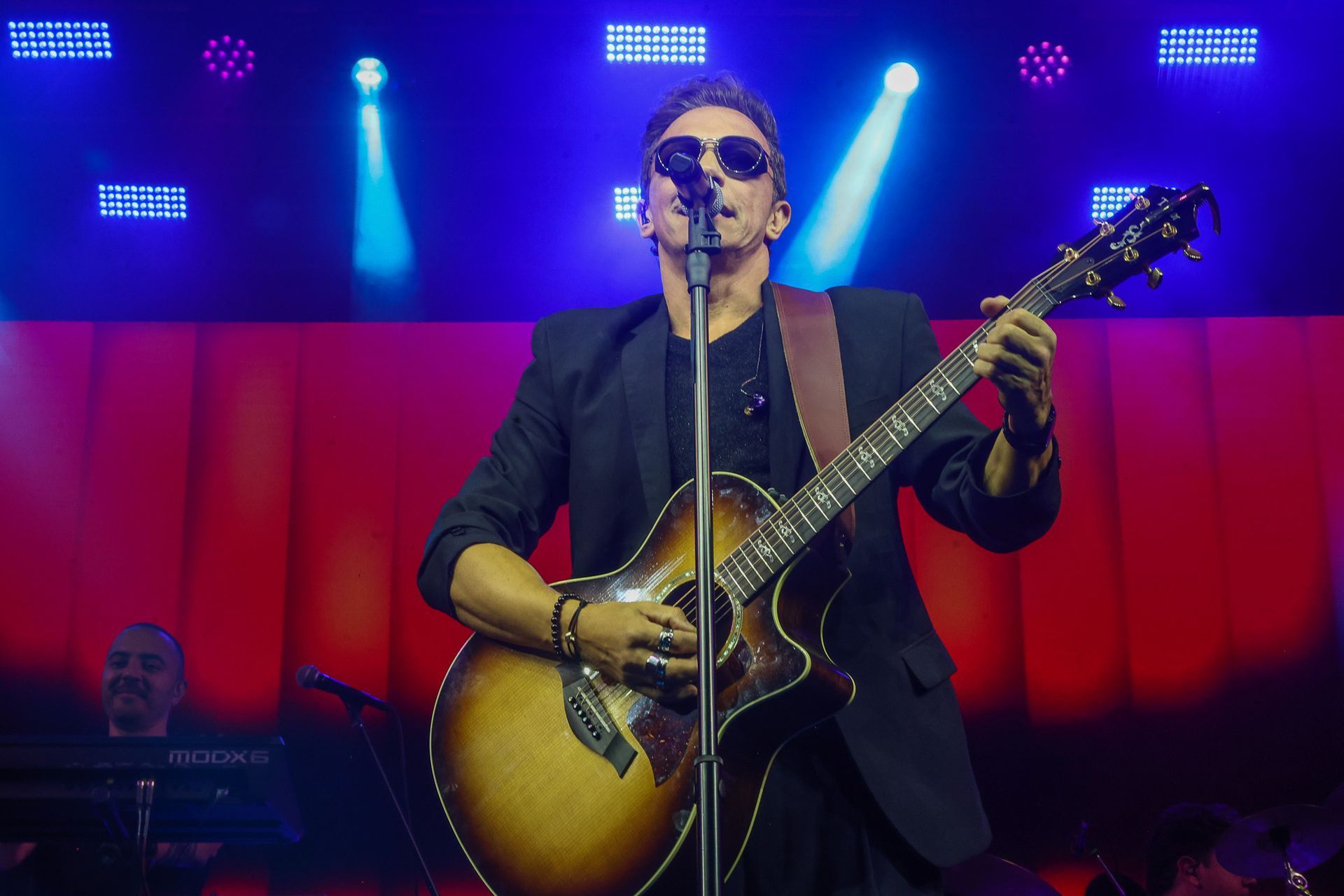 Um músico usando óculos escuros toca violão e canta em um microfone em um palco com luzes vermelhas.